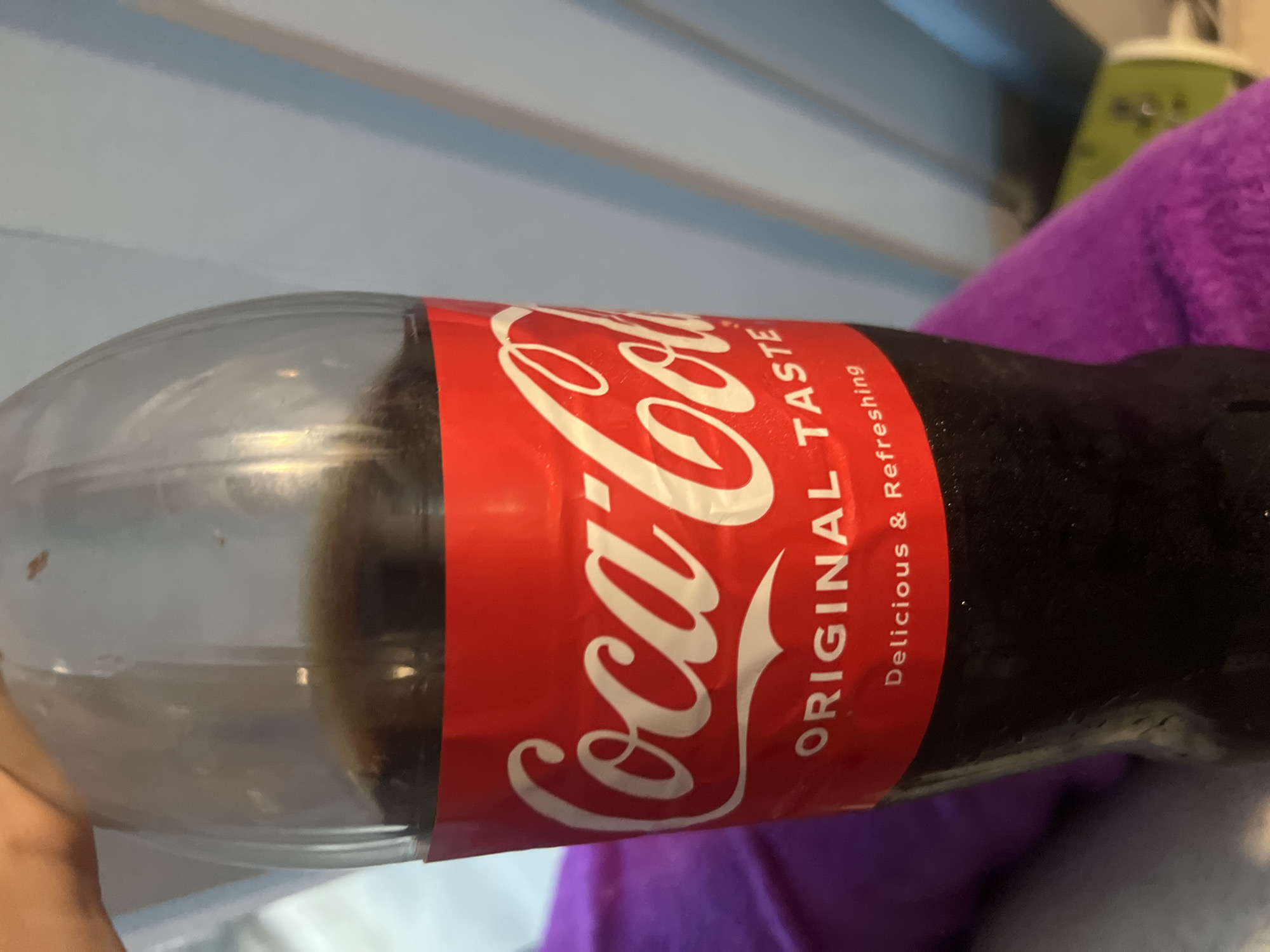 A close-up, vertical shot shows a clear plastic bottle of Coca-Cola. The label is red with white lettering. The top of the label displays the Coca-Cola logo, which is white and stylized. Below the logo, the words "ORIGINAL TASTE" are written in white. Underneath that, the words "Delicious & Refreshing" are also written in white. The bottle is partially filled with a dark brown liquid, presumably Coca-Cola. The bottle appears to be held by a hand, which is partially visible at the top of the frame. The background is out of focus and appears to be a light blue and white striped surface, possibly a wall or a piece of furniture. The overall lighting is soft, with some reflections visible on the bottle.