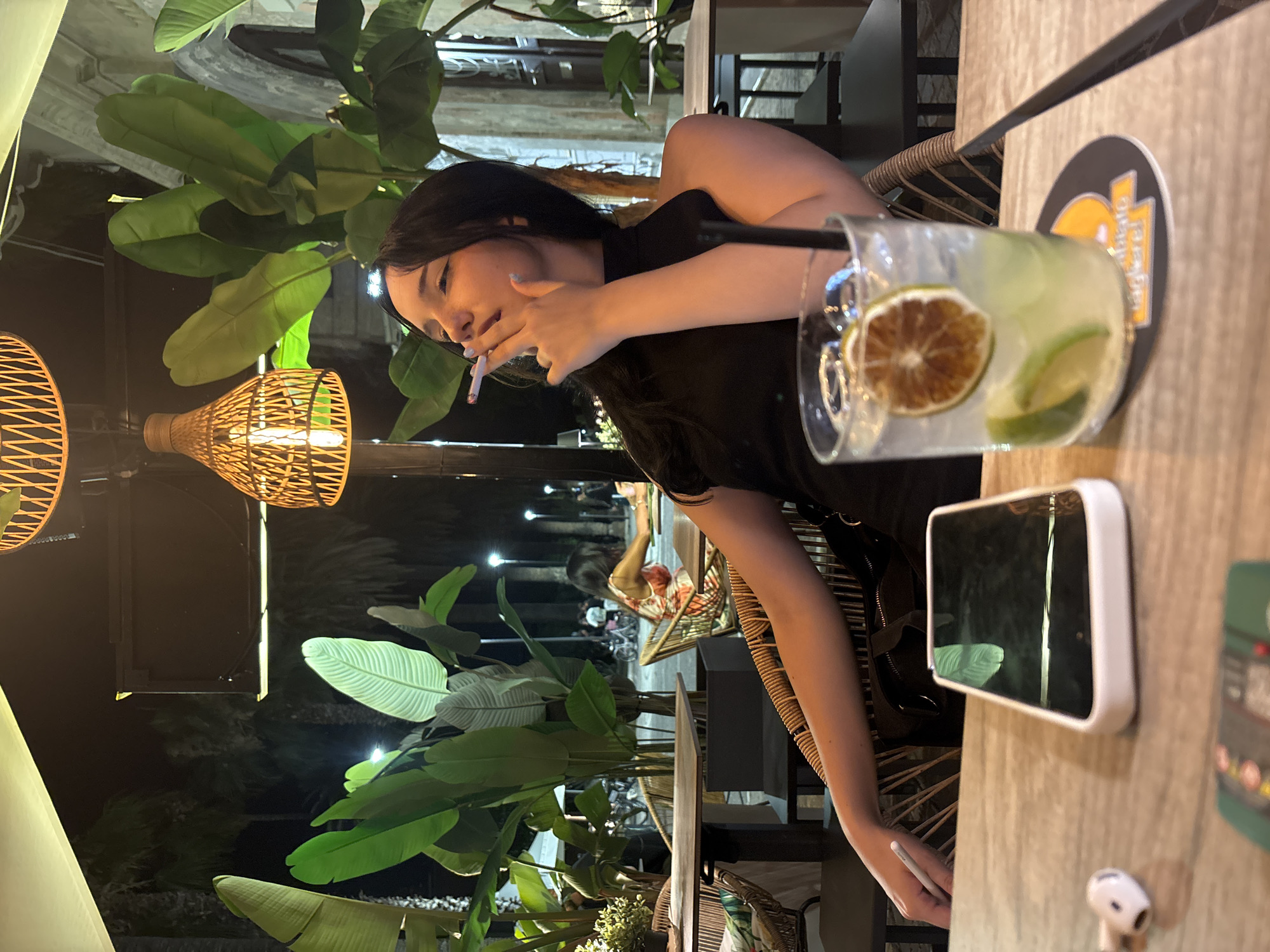 A young woman with dark hair is smoking a cigarette while sitting at an outdoor cafe in San Benedetto del Tronto, Italy. She is wearing a black top and is seated in a wicker chair. In front of her is a glass of drink with ice, lime, and a dried citrus slice. A smartphone and a pair of wireless earbuds are on the table. In the background, palm trees and other vegetation are visible, along with ambient lighting and other patrons at tables. The setting appears to be at night.