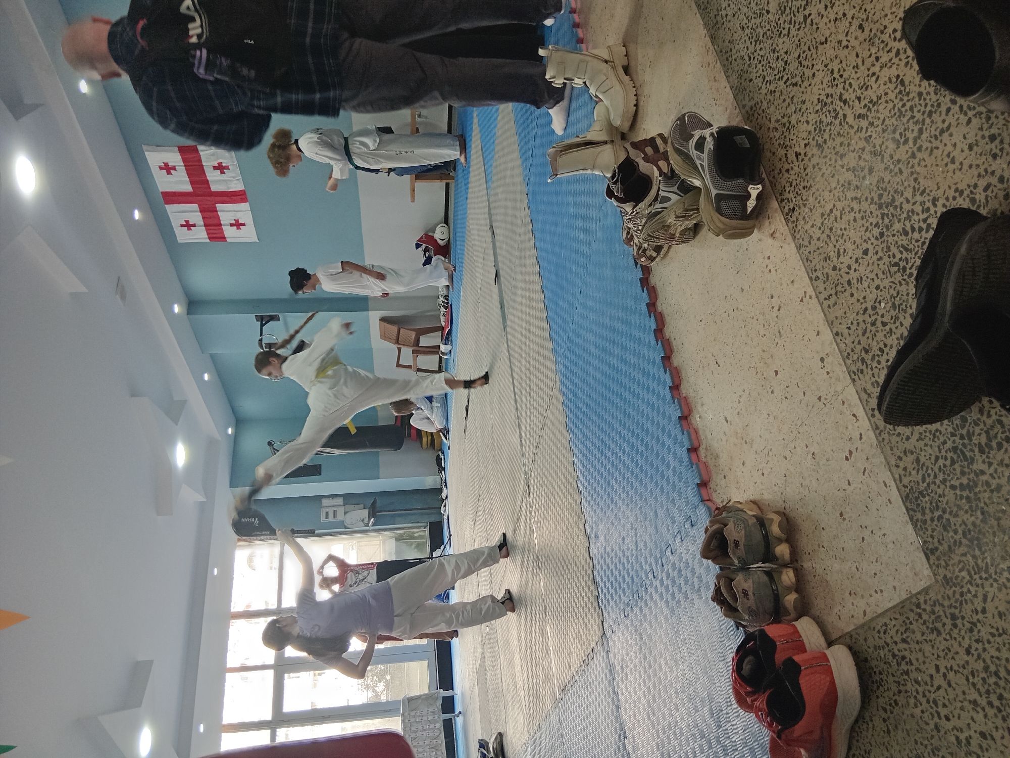 The photograph depicts an indoor martial arts training space. Several individuals wearing traditional white uniforms are practicing techniques. One person is performing a kick, while another is holding a target pad. A Georgian flag hangs on the wall. There are spare shoes piled up to the side.
While it is difficult to ascertain the exact location from this photograph alone, considering the flag and the question context, the location is presumably in the city of Batumi, Georgia.
