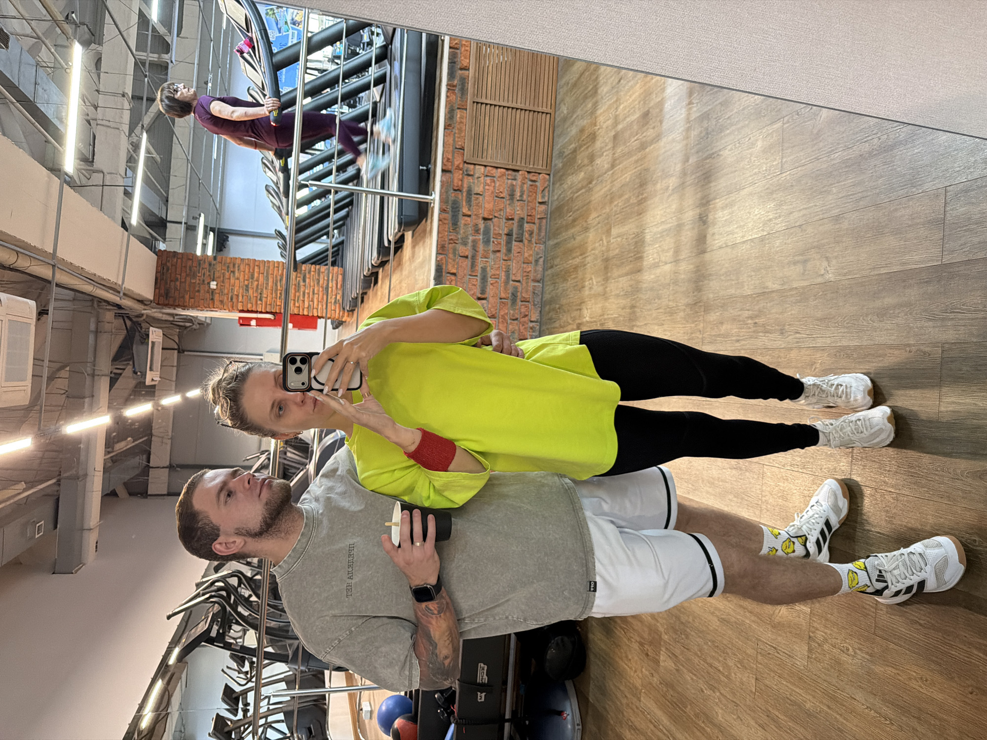 A man and a woman stand side-by-side taking a selfie in a mirror. The man has a beard, light brown hair and wears a gray t-shirt, white shorts with black trim, and white Adidas shoes with lemon-themed socks. He holds a black cup. The woman wears a bright yellow shirt, black leggings, and white sneakers. She holds the phone up to take the picture. Behind them are exercise machines in a gym. The scene is located in Chaikovsky, Russia. Another person can be seen working out in the mirror's reflection.