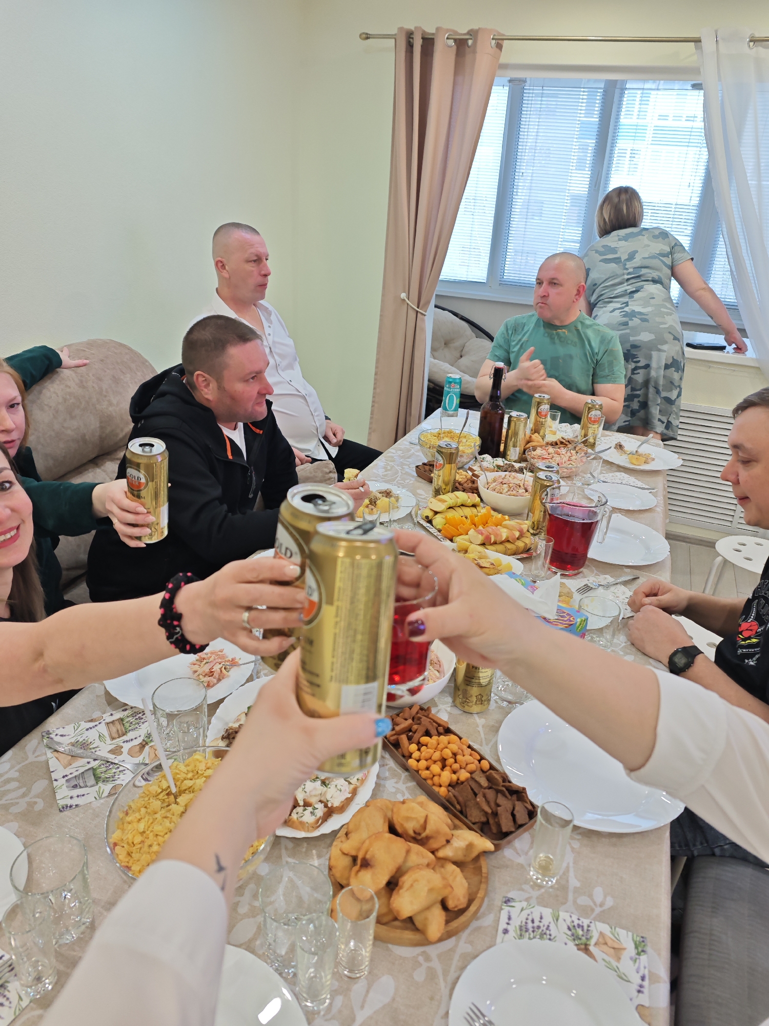 An indoor social gathering, likely a celebratory meal or party, is depicted with at least seven individuals gathered around a dining table. The scene is set in what appears to be a home or apartment in Markovskiy, Russia, characterized by light-colored walls and curtains on a window fitted with horizontal blinds.
The long table is covered with a patterned tablecloth featuring lavender and garden tool motifs. It is laden with a variety of food items: a large bowl of yellow chips or crisps, a tray of dark crackers and peanuts, small fried pastries, salads, cut fruit (apples and oranges), and open-faced sandwiches or canapés. Beverages include several gold-colored beer cans, one dark glass bottle, and a pitcher of red juice or punch.
In the foreground, several hands are engaged in a toast, clinking gold beer cans together. One can prominently displays the text "GOLD PREMIUM," and another has "Premium" visible. A woman's hands in the foreground, wearing a ring and blue nail polish, hold a can while another person's hand holds a can with "POLSKA" visible.
Guests are seated around the table and on a nearby sofa. Two men with shaved heads are on the sofa; one in a white shirt, another in a black hoodie. A man in a green t-shirt sits at the table, engaged in conversation. Another man in a black t-shirt is visible on the right, wearing a watch. A woman in a camouflage-patterned dress stands near the window with her back to the camera. Another woman with long hair, wearing a green top, is seated on the sofa, also holding a beer can.
The atmosphere appears casual and convivial, with people socializing and celebrating. The time of day, while indoors, suggests daytime or early evening due to the natural light filtering through the window blinds.