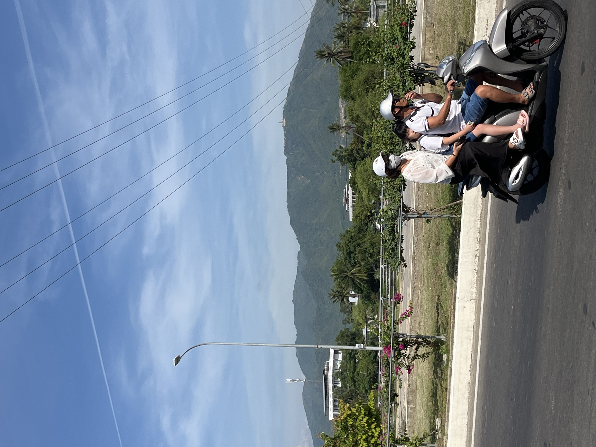 Three people are riding on a scooter on a paved road. The scooter is silver and is moving from left to right. The person in the back is wearing a white shirt, black pants, and a white helmet with a face mask. The person in the middle is a child wearing a white shirt and blue shorts, and is sitting between the other two. The person in the front is wearing a white polo shirt, blue shorts, and a silver helmet. They appear to be driving and possibly smoking. The road is asphalt and there is a white concrete barrier on the left side. In the background, there is lush green vegetation, trees, and buildings. In the far background, there are mountains under a bright blue sky with scattered clouds and power lines. The overall scene suggests a warm, sunny day. This scene is located in Nam Nha Trang Ward, Vietnam.