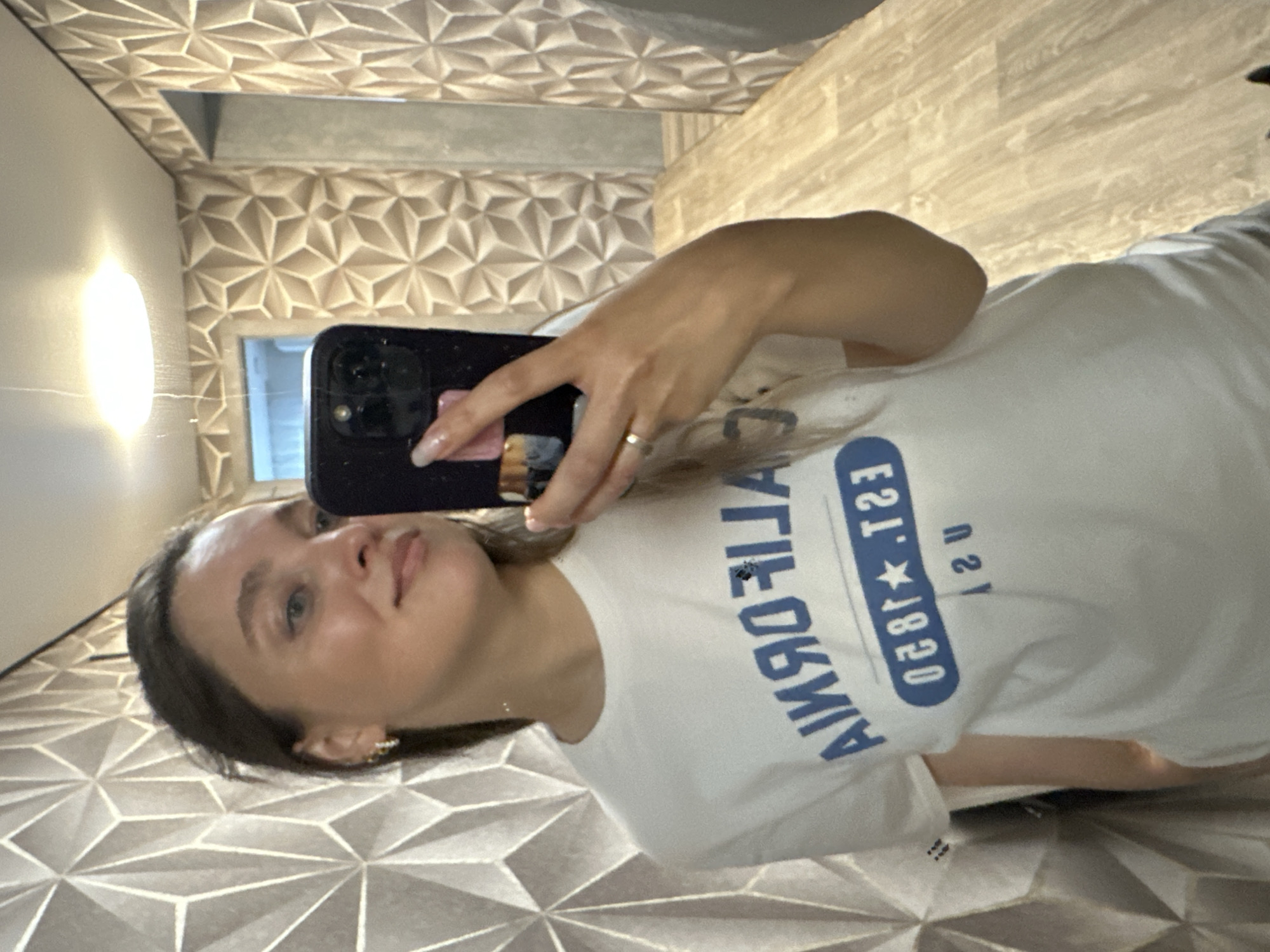 A woman takes a mirror selfie in a hallway. She is wearing a white t-shirt with blue lettering that says "CALIFORNIA EST. 1920 USA." The hallway has textured geometric wallpaper and a wood-look floor. The woman has long brown hair and is wearing earrings and a ring. She is holding a black smartphone to take the picture.