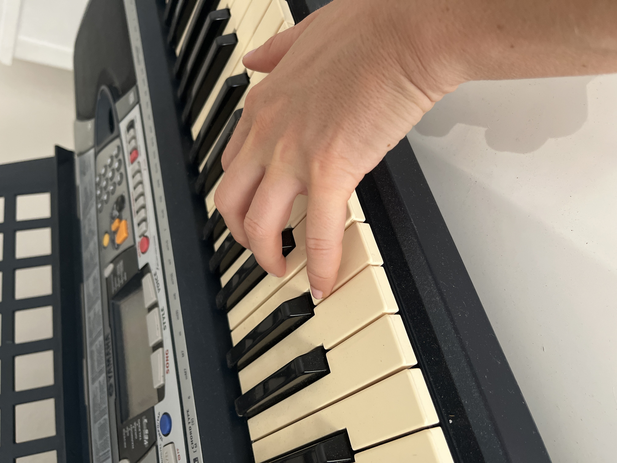 An overhead shot shows a person's hand playing a black and white keyboard. The keyboard is on a white surface, possibly a table. The background is a blurry white wall and a black music stand. There are no discernible landmarks to indicate the city of Athens, Greece.
