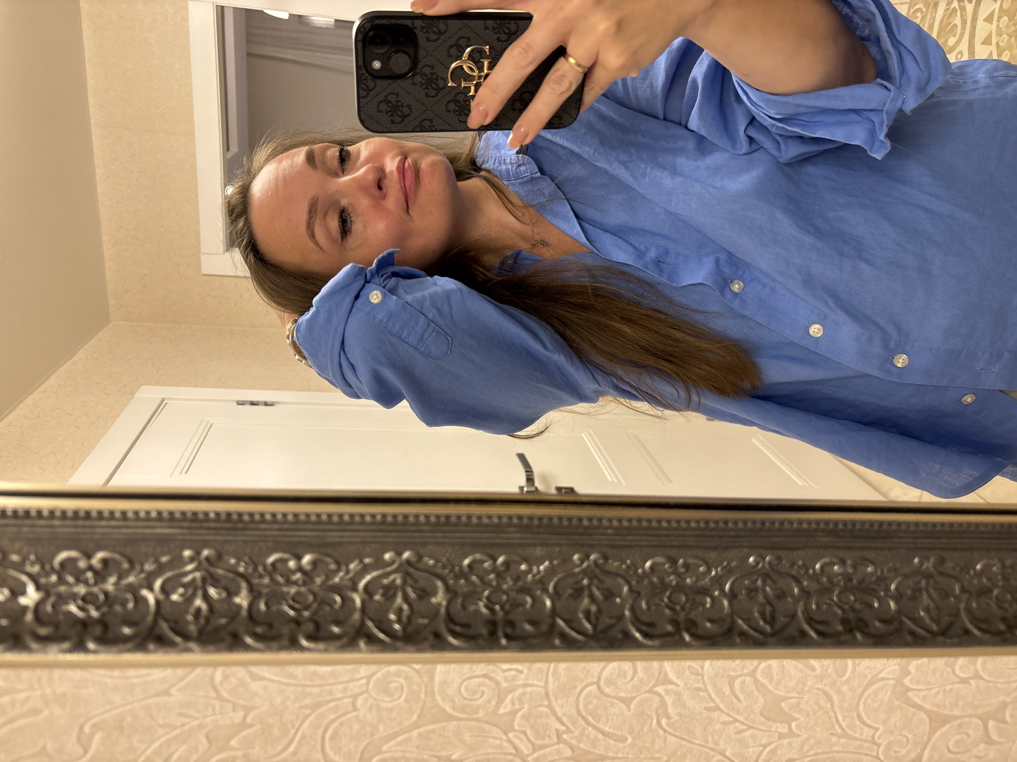A woman is taking a selfie in a mirror. She is wearing a blue button-down shirt and has her hair in her hand. The mirror has an ornate frame. The background shows a doorway and patterned wallpaper. The location is not discernible from the image.