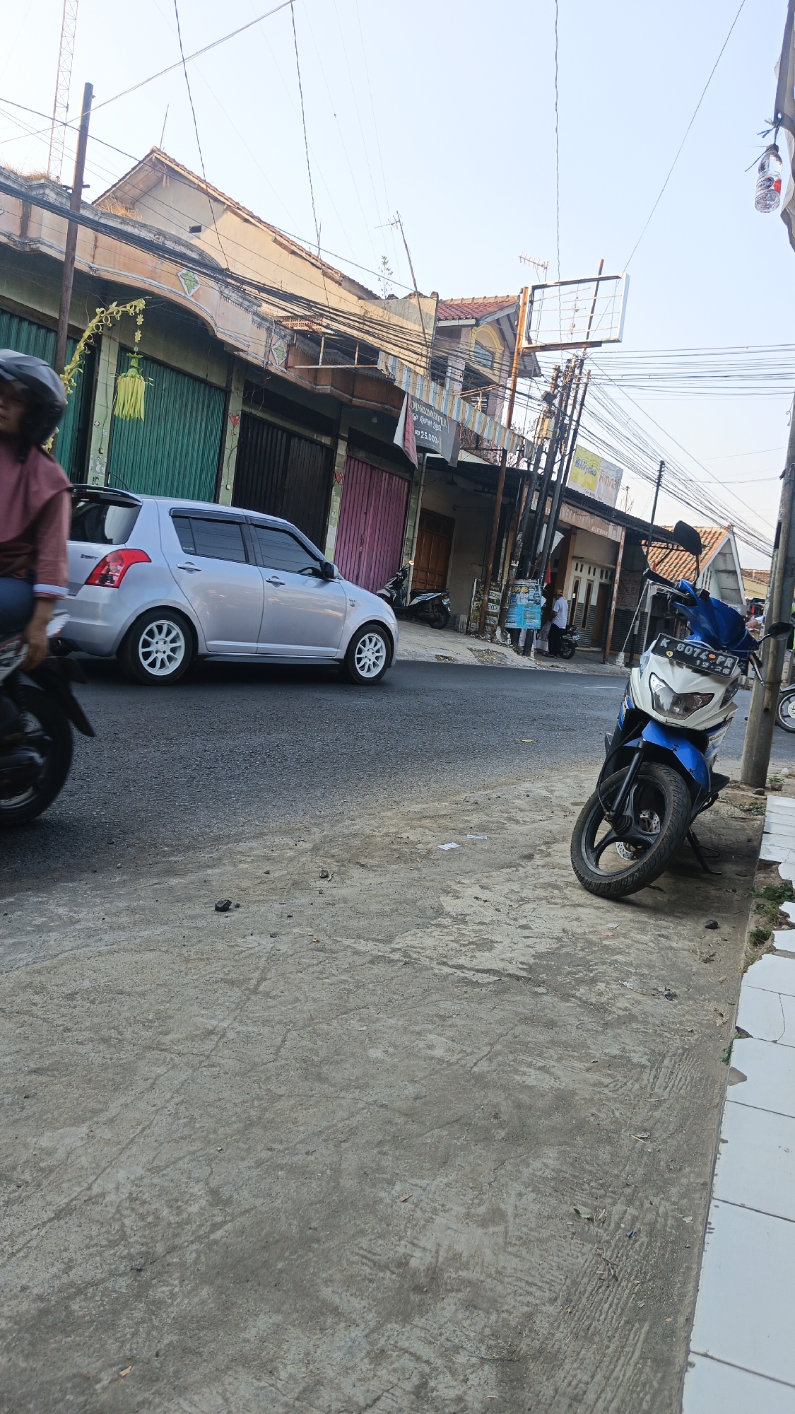 The image shows a street scene in the city of Dawe, Indonesia. In the foreground, a blue and white motorcycle is parked on the side of the road. The motorcycle has a license plate that reads "K 6074 PR". Further down the street, a silver car is driving on the road. The car is a hatchback with white rims. A person on a motorcycle is partially visible on the left side of the image, wearing a pink top and a helmet. The background consists of buildings with colorful shutters and overhead power lines. The sky is clear and blue. The scene appears to be a typical street in a populated area, with vehicles and pedestrian activity.