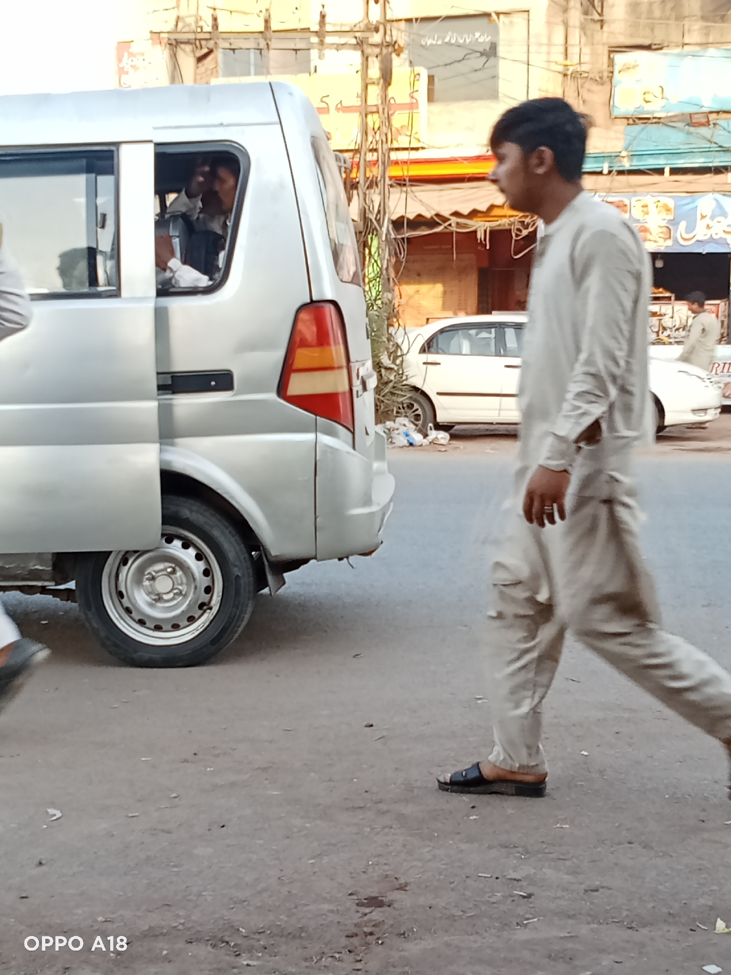 A medium shot captures a man walking on a street in an urban environment. The man is dressed in a light-colored shalwar kameez and wears black sandals. He is walking from right to left in the frame, positioned in front of a silver van that has its sliding door open. A man is partially visible inside the van. In the background, a white car is parked alongside the road, and another man is visible near the car. The urban setting includes buildings with signs in a language other than English, power lines, and storefronts. The street surface is asphalt. "OPPO A18" is printed in the bottom left corner of the image.