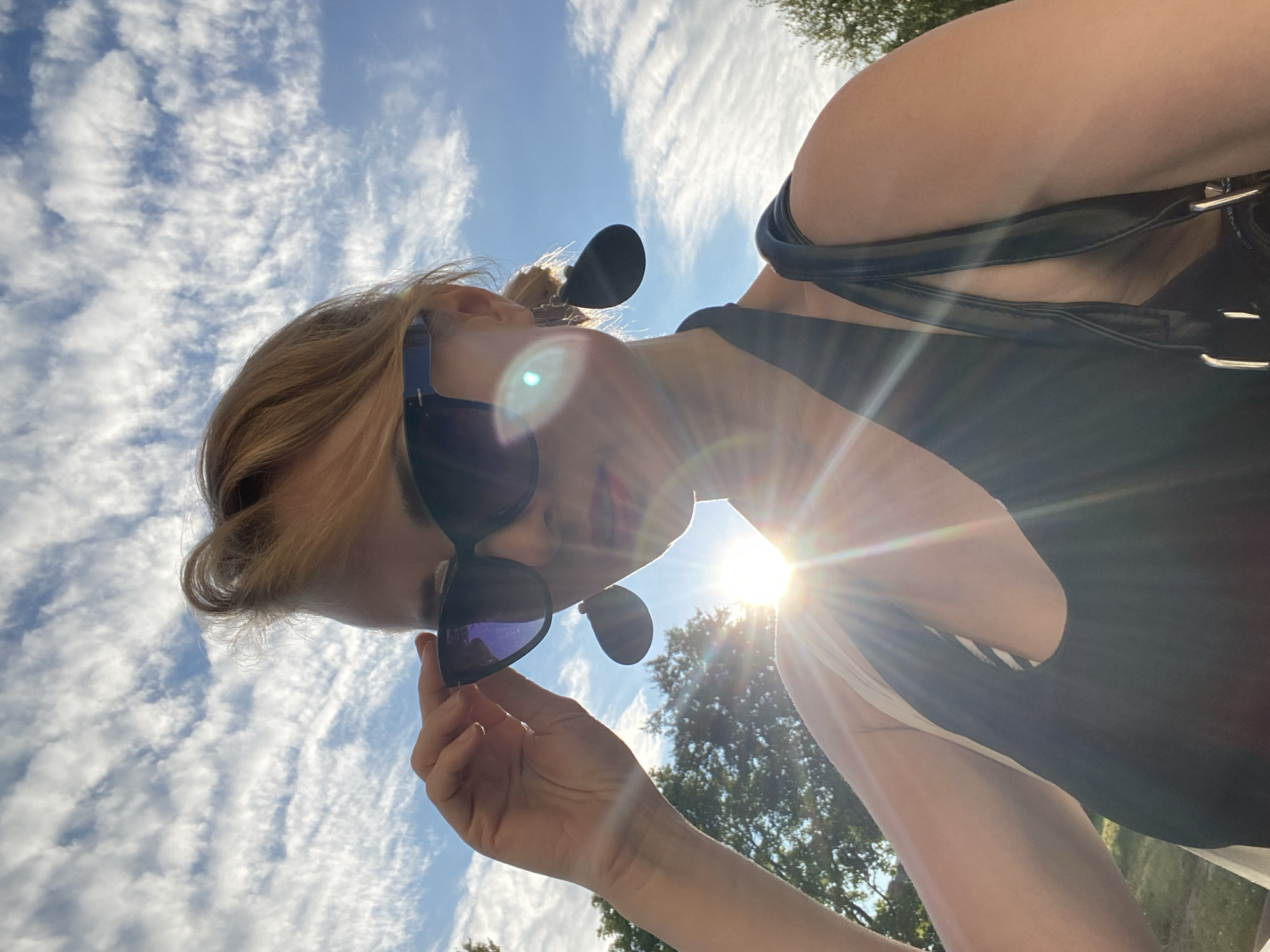 A woman is taking a selfie outdoors, looking up at the camera. She is wearing large, dark sunglasses and large, black, teardrop-shaped earrings. Her hair is blonde and styled with volume at the crown. She is wearing a black halter top. Her right hand is raised, touching the brim of her sunglasses. The sun is shining brightly, creating a lens flare that emanates from the lower right of the frame. The sky is blue with scattered, wispy white clouds. Green trees are visible in the background to the left and right. The ground appears to be grass, suggesting a park or outdoor recreational area. The context of Lida, Belarus, implies this is a public outdoor space within that city.