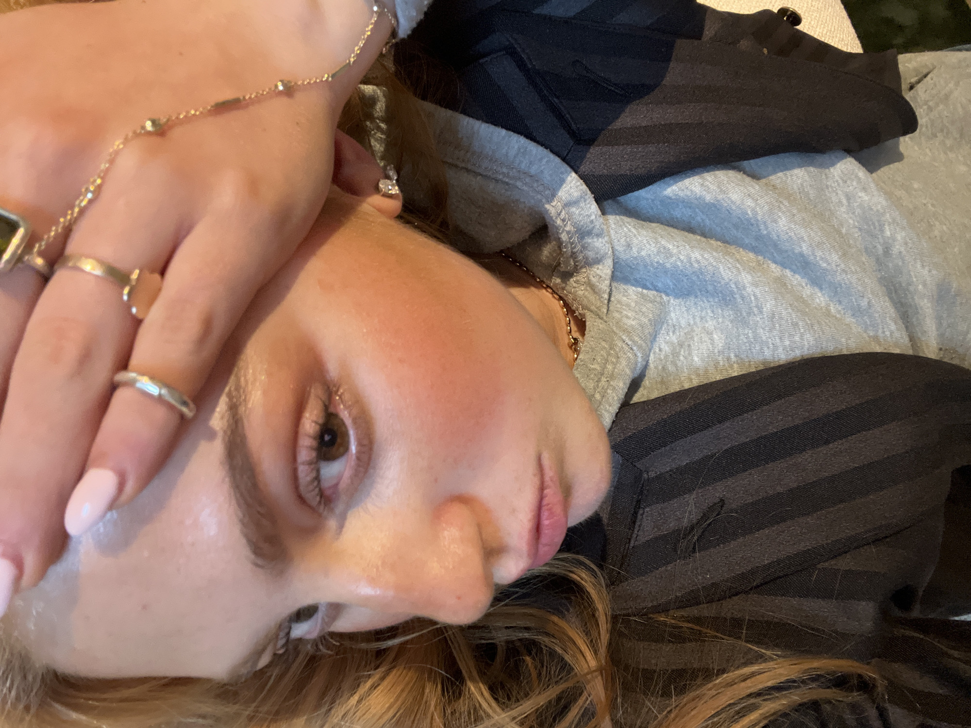 A young woman is lying down, her head tilted to the side. Her left hand is raised near her forehead, adorned with multiple rings. She has light skin, brown eyes, and light brown hair. She is wearing a gray hooded sweatshirt layered under a dark striped shirt. The background is indistinct. The image appears to be taken indoors. There is no visual information to suggest the location is Paris, France.