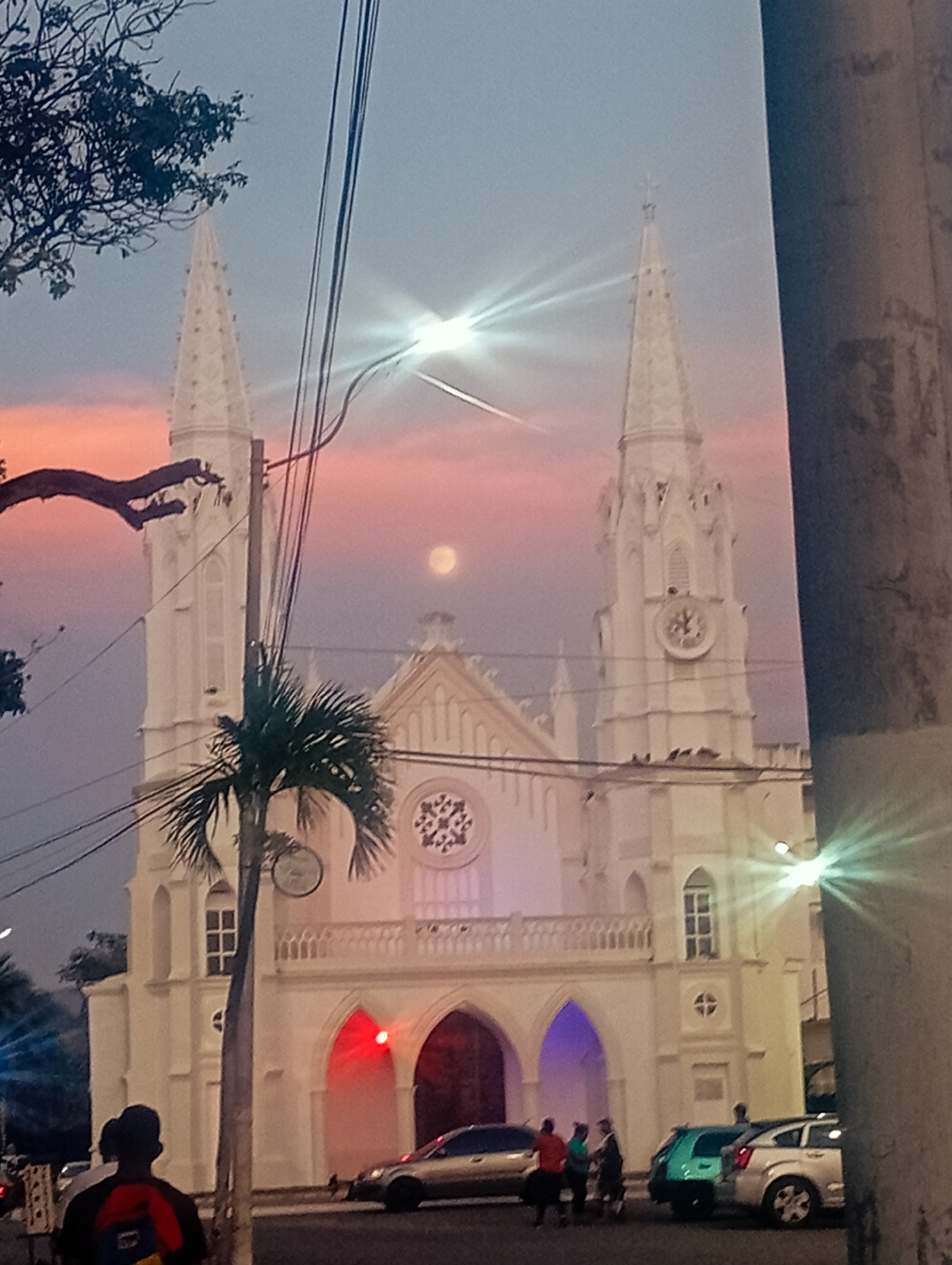 This image captures an evening scene in Juan Griego, Venezuela, centered around a grand white Gothic-style church, likely the Santa Rosa de Lima Church. The church features two prominent spires, ornate arched entrances, and intricate architectural details, including a clock face on the right spire and a circular rose window-like element above the main entrance. The primary entrance arches are distinctively illuminated with colored lights, red on the left and purple on the right, enhancing the twilight ambiance.
The sky behind the church showcases a beautiful sunset, with a gradient of pink, orange, and purple fading into a lighter blue-grey. A bright, full moon is clearly visible in the sky, positioned to the left of the right spire. Dominating the foreground and midground are numerous dark power lines strung across utility poles, some partially obscuring the church. A bright streetlight on the right creates a distinct lens flare.
On the street in front of the church, several people and vehicles are present. A person with a red and blue backpack is seen from behind in the lower left. A small group of individuals is gathered near parked cars, which include a silver sedan, a light green hatchback, and a white SUV. Lush foliage, including a large tree branch on the upper left and a palm tree in the mid-left, adds to the tropical setting.
The scene captures a peaceful evening, blending architectural beauty with natural elements like the sunset and moon, set against the backdrop of an active urban environment. No legible text is visible in the media.