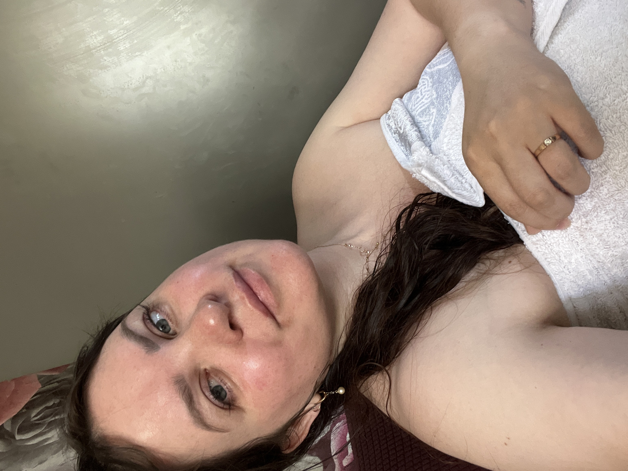 The image shows a medium shot of a young woman wrapped in a white towel, taken indoors. The woman has dark brown hair that is wet and falls around her shoulders. She is looking towards the camera with a slight smile. She is wearing a delicate gold necklace and a gold earring with a pearl. On her left hand, she is wearing a gold ring with a small diamond. The background is a wall with a textured, possibly painted, surface. The lighting is soft, creating a warm and intimate atmosphere. The image does not provide any information about the specific location in the city of Noviy, Russia.