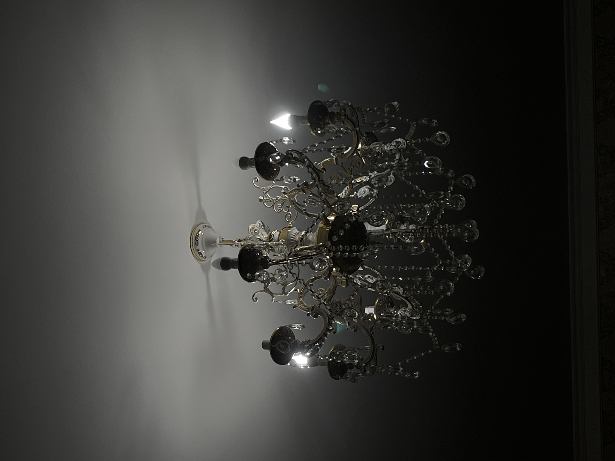 A grand, ornate chandelier with multiple arms and crystal accents hangs from a white ceiling. The chandelier has a brass-colored frame and appears to be lit by several bulbs, some of which emit a warm, bright light, while others are darker. The ceiling is evenly lit, with subtle shadows cast by the chandelier's structure. The lower portion of the image transitions to a very dark, almost black background, obscuring whatever lies beneath. No people are visible in the scene. The image does not provide any information about its location in the city of Qūryq, Kazakhstan.