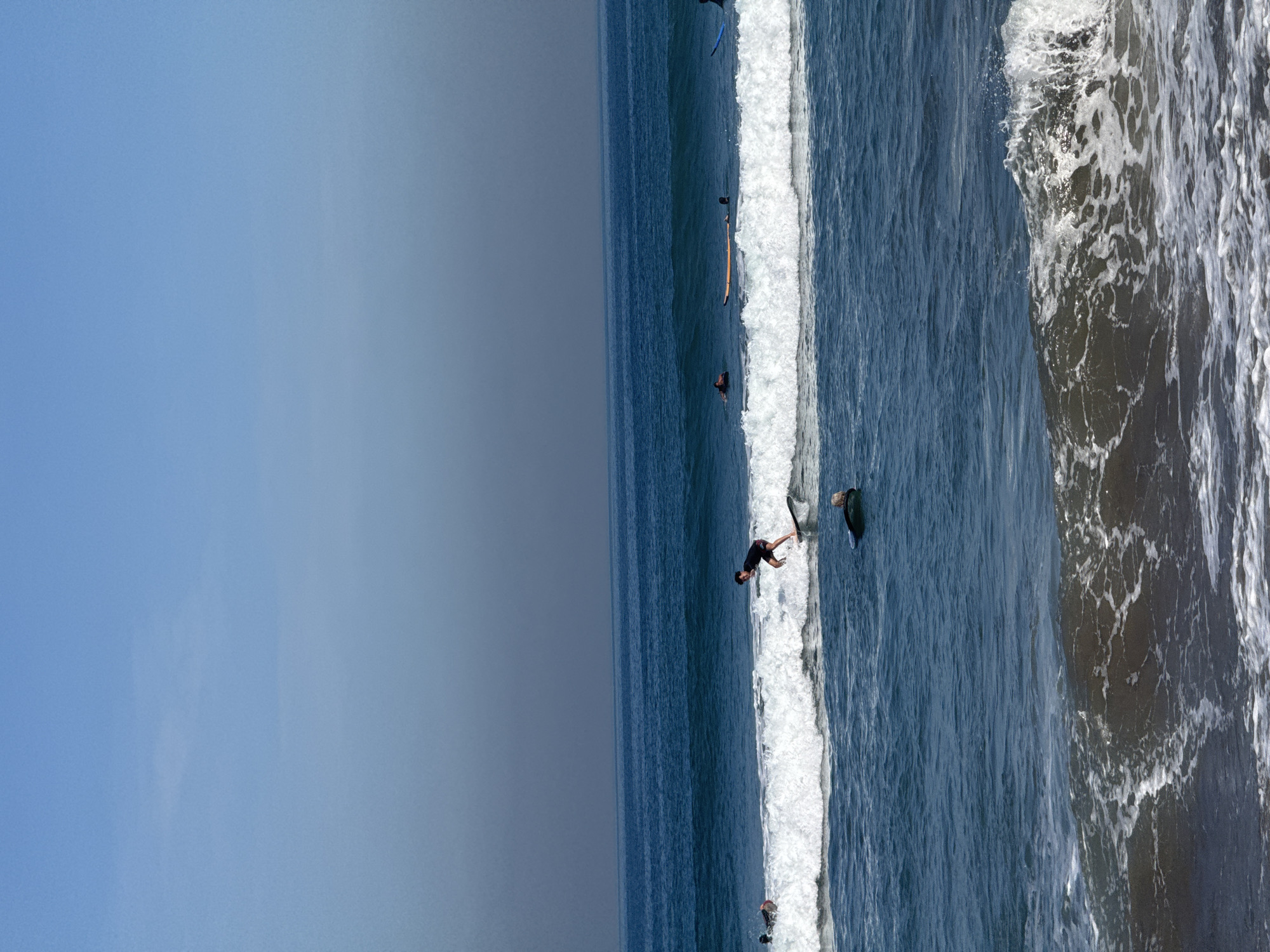 In Kuta, Indonesia, on a clear day with a hazy blue sky, several surfers are visible in the ocean. The primary subject is a male surfer in the foreground, actively riding a wave on a surfboard, with his body positioned in a crouching stance. Another surfer is partially submerged in the water to the left of the main surfer, with only their head and shoulders visible. In the middle ground, several other surfers are scattered across the water, some on their boards, others in the process of paddling or waiting for waves. The ocean displays blue water with white-capped waves, indicating moderate surf conditions. The scene is characterized by the natural environment of a beach and ocean, with no artificial structures immediately visible.