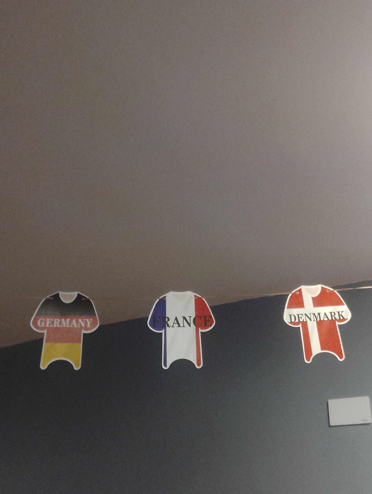 Three t-shirt shaped cutouts with national flags are hanging on a dark wall. From left to right, the cutouts represent Germany, France, and Denmark. The German cutout has the colors black, red, and yellow, and the text "GERMANY" is written across it. The French cutout has the colors blue, white, and red, and the text "FRANCE" is written across it. The Danish cutout has the colors red and white, and the text "DENMARK" is written across it. The cutouts are hanging at different heights and are positioned in a horizontal line. To the right of the Danish cutout, there is a small, light gray rectangular object attached to the wall. The background is a plain, dark gray wall.