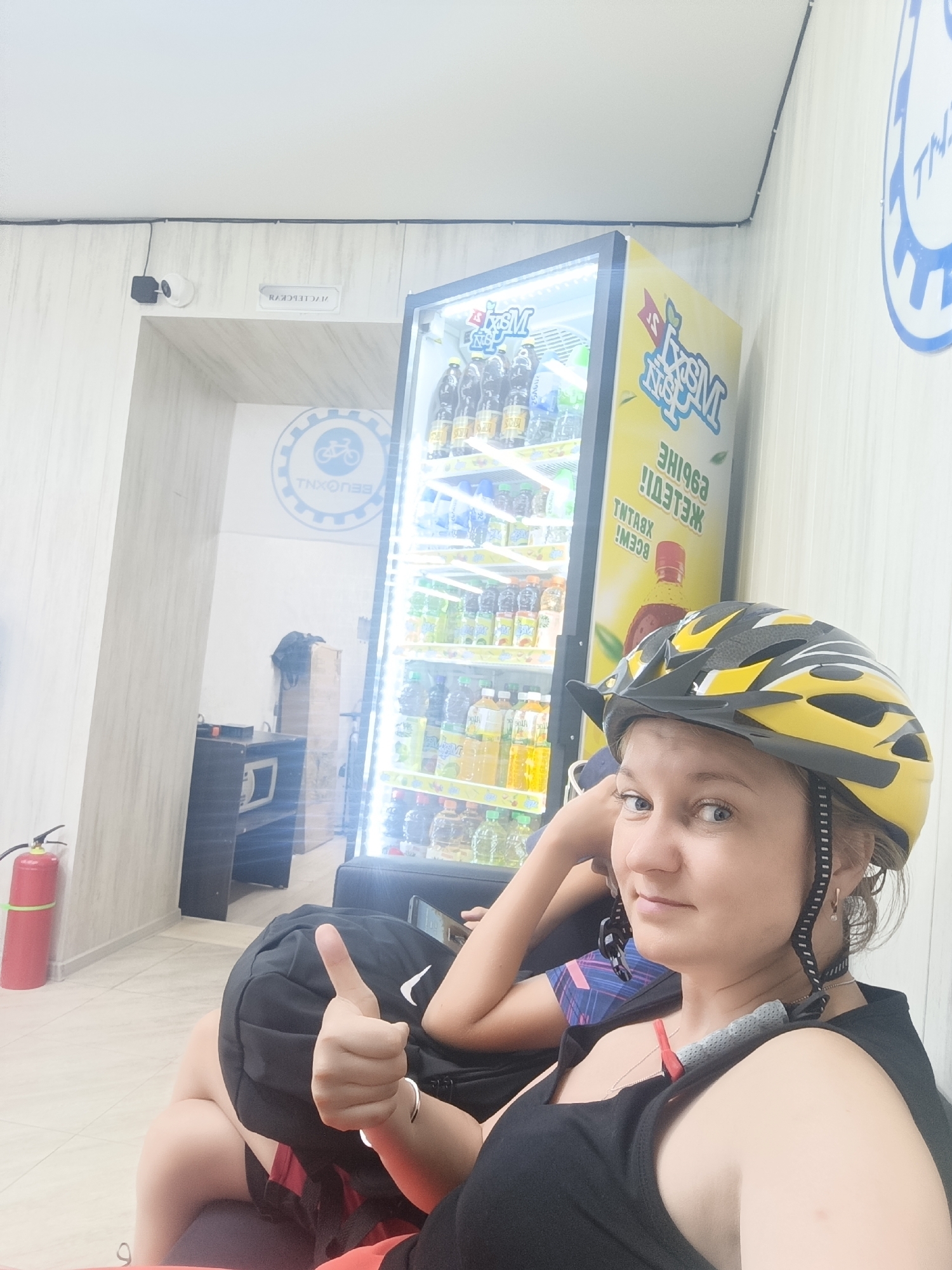 A woman wearing a yellow and black bicycle helmet is sitting in front of a brightly lit vending machine stocked with bottled beverages. She is wearing a black tank top and has a silver necklace. She is giving a thumbs-up gesture. Behind her, a red fire extinguisher is visible leaning against a wall. The vending machine has yellow branding with Russian text that reads "ТНХОПББ" (TXNOPBB) and "ВСЕМИ" (VSEMI). The environment appears to be indoors, possibly a rest stop or a shop, given the presence of the vending machine and the bicycle-themed logo on the wall. The overall setting is clean and well-lit.