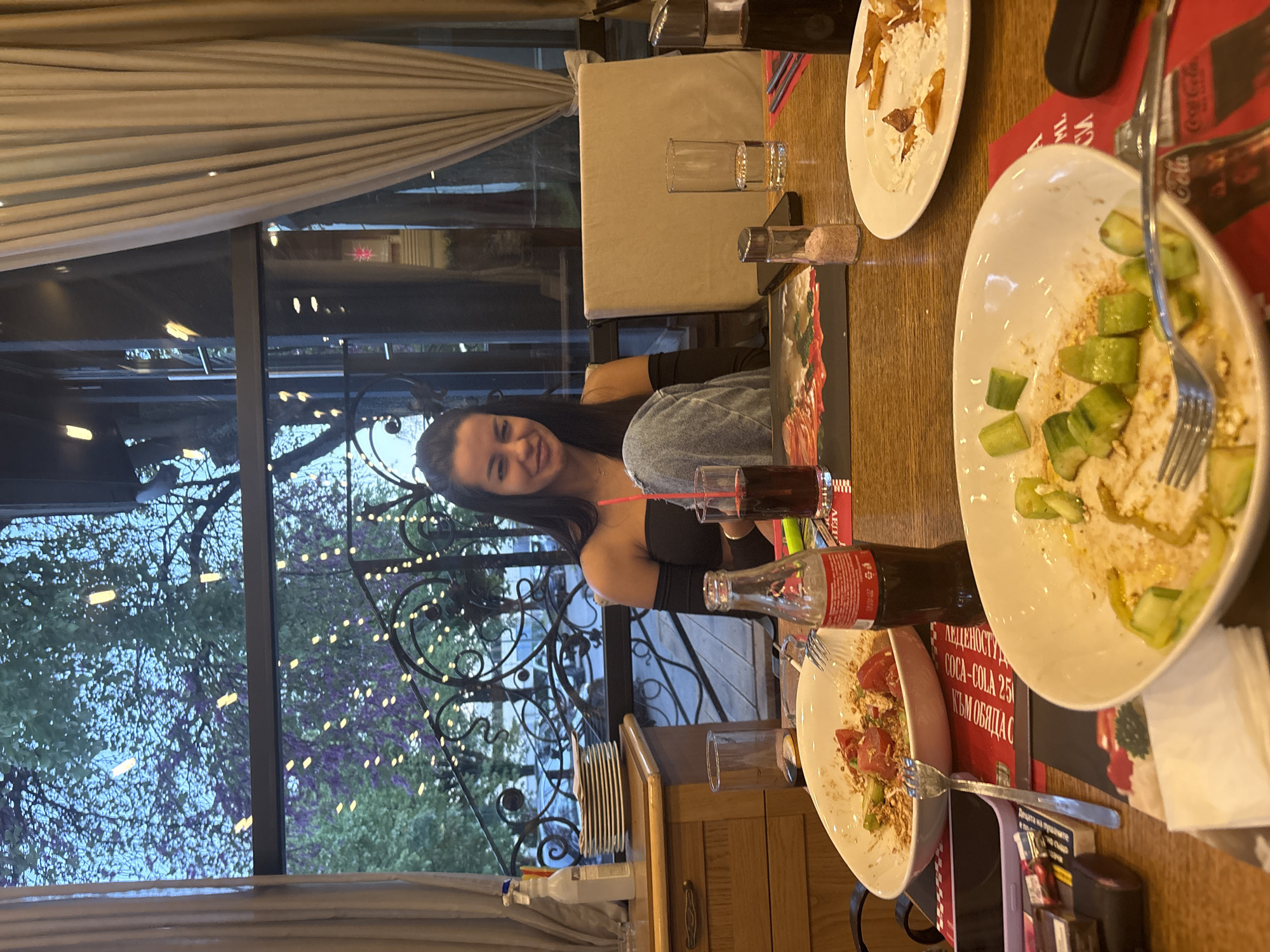 The image shows a young woman sitting at a table in what appears to be a restaurant or cafe in Plovdiv, Bulgaria. She is smiling at the camera. On the table are several plates with food remnants, a Coca-Cola bottle, a glass of liquid with a straw, a salt shaker, and a cell phone. The table is set with a red Coca-Cola placemat. Outside the window behind her is a view of trees, wrought iron decor, and possible building lights, suggesting it's either dusk or evening. The presence of empty plates indicates a meal has just been completed.