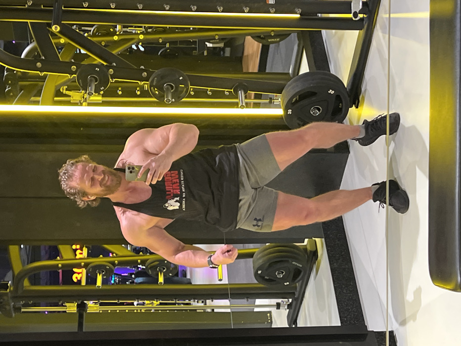 An upright full-body mirror selfie captures a muscular man inside a modern gym in Saint Petersburg, Russia. The man, with curly brown hair and a beard, stands smiling at the camera, holding a light-colored smartphone in his left hand to take the picture. He wears a black tank top, grey athletic shorts, and black athletic shoes, with a dark smartwatch visible on his left wrist.
The black tank top features a mirrored white text and a graphic of a strongman or gorilla. The mirrored text clearly reads "MEVK KEMDITU," with smaller mirrored text underneath that appears to say "CALLED KLAVEV OV VENEGY." His grey shorts display the Under Armour logo on the left thigh.
Behind the man, various pieces of gym equipment are visible, including black weight plates marked with the number "30" on a barbell. The gym's interior is brightly lit by prominent yellow or gold strip lighting integrated into the horizontal structures of the equipment, creating a sleek and modern ambiance. The setting suggests a well-maintained fitness facility.
