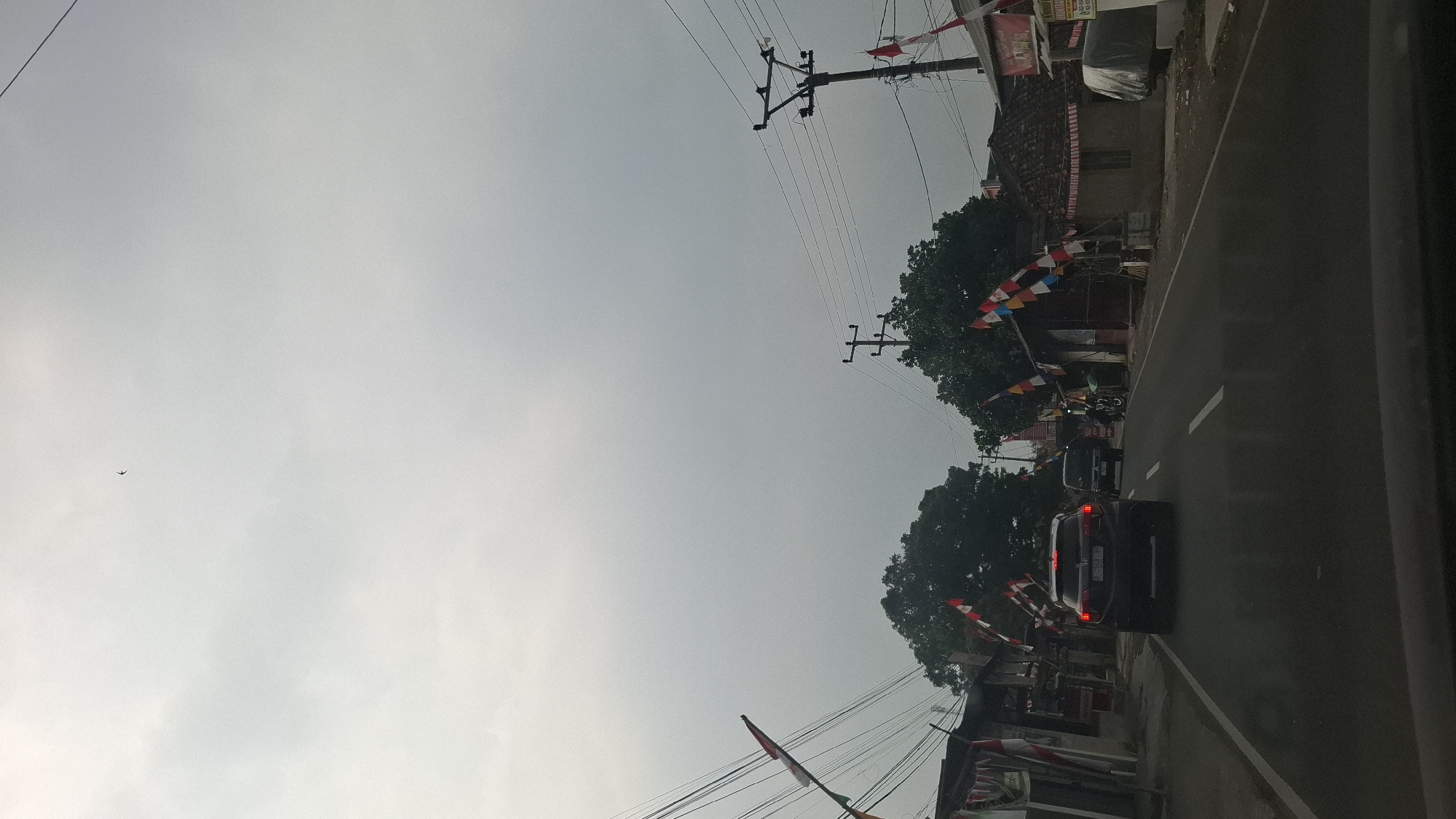 The image depicts a street scene in Peuteuycondong, Indonesia, under an overcast sky. A dark-colored SUV is traveling on a paved road, followed by a dark van. The road is lined with buildings and trees, with numerous Indonesian flags displayed along the roadside, suggesting a festive or commemorative occasion. Power lines and utility poles are visible overhead. The overall atmosphere is subdued due to the cloudy weather.