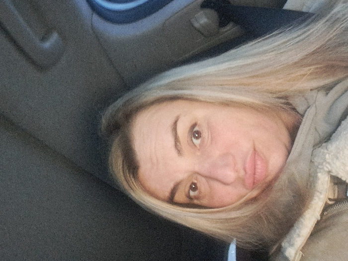A woman with long blonde hair gazes at the camera with a slight smile. She wears a beige jacket with a fur-lined collar. The background appears to be the interior of a car, with a dark gray panel behind her head. The image is taken at an angle, giving a slightly tilted perspective of her face. Her eyes are brown, and her skin is fair.