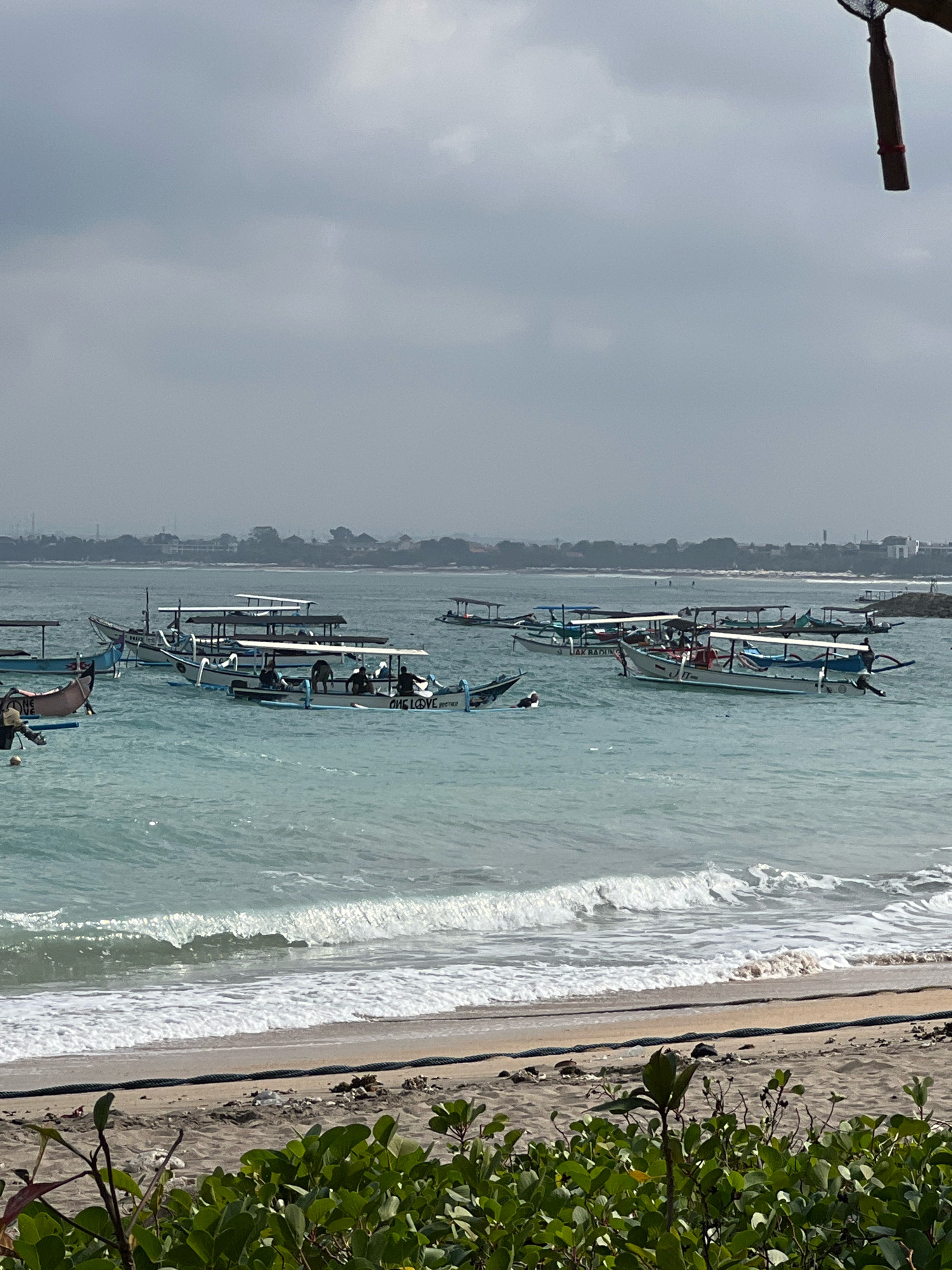 The image depicts a cloudy day at Kuta Beach, Bali, Indonesia. Several traditional Balinese fishing boats, known as jukung, are anchored in the turquoise water. Some of these boats have canopies providing shade, and passengers can be seen in some of them. The waves are breaking gently on the sandy shore in the foreground, where some green foliage is visible. In the distance, the coastline is lined with buildings and lush greenery. A makeshift fishing net is also visible in the upper right corner of the image. The scene suggests a calm, everyday moment at a popular tourist destination.