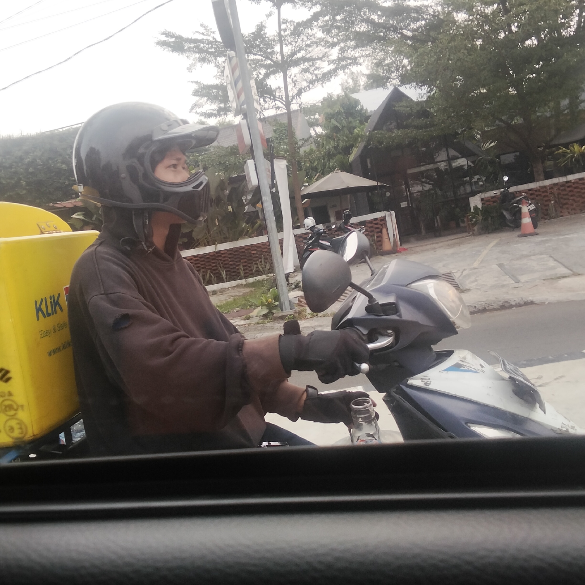 The image captures a person on a motorcycle in an outdoor urban setting, possibly a street in Bogor, Indonesia, given the context. The person, wearing a black helmet and a brown hooded jacket with a torn sleeve, is positioned on the left side of the frame, facing to the right. They are holding the motorcycle handlebars with gloved hands and reaching for a clear plastic bottle from what appears to be a yellow delivery box attached to the motorcycle. The delivery box has the word "Klik" printed on it. The background features trees, buildings with dark roofs, and a brick wall. Two other motorcycles are visible in the distance. The overall lighting suggests an overcast day.