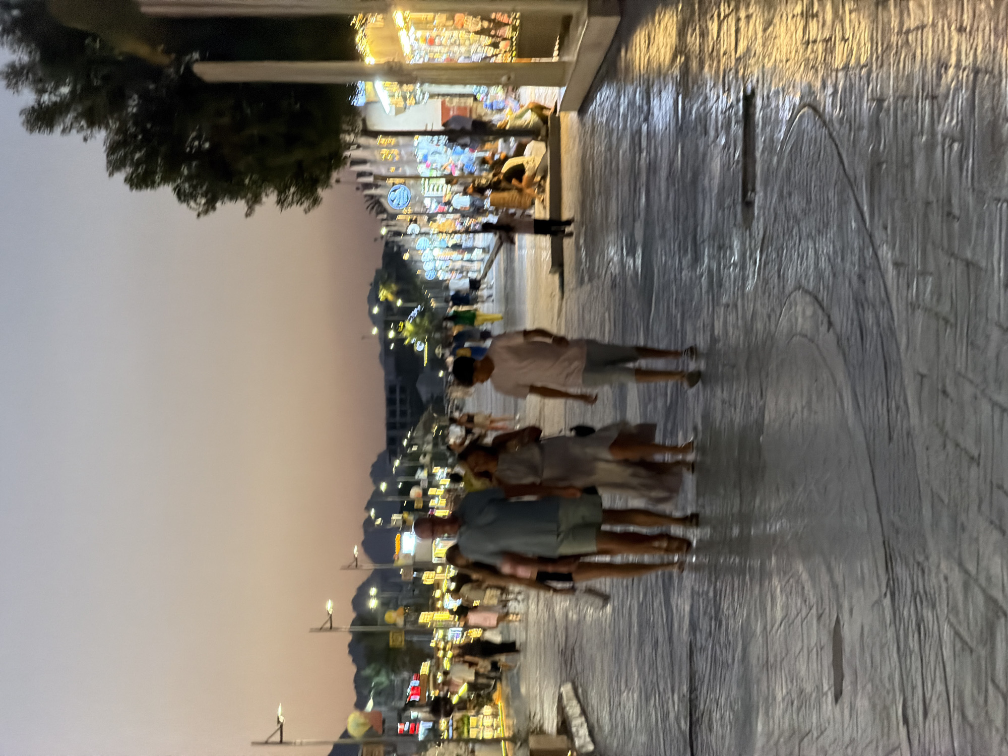 The image depicts a dusk scene on a pedestrian walkway in Sharm El Sheikh, Egypt, likely along the Naama Bay promenade. The sky is a soft gradient of pale blue and orange. The walkway is paved with textured grey stone, wet and reflecting the lights from storefronts and street lamps.
In the foreground, a family is walking away from the camera. A man in a blue polo shirt and light shorts is in the center, flanked by a woman in a long, light-colored dress on his right and another woman in a lighter dress and dark shorts on his left. A young boy in a pink t-shirt and shorts is walking ahead of them. Further down the walkway, other pedestrians are visible, some browsing storefronts.
The buildings lining the walkway are adorned with numerous lights, creating a festive atmosphere. The storefronts appear to be shops selling various goods, some with colorful displays. Street lamps with angled fixtures are also present, illuminating the path. In the background, mountains are silhouetted against the fading light, and palm trees add to the tropical ambiance. The overall impression is one of a lively evening stroll in a tourist area.