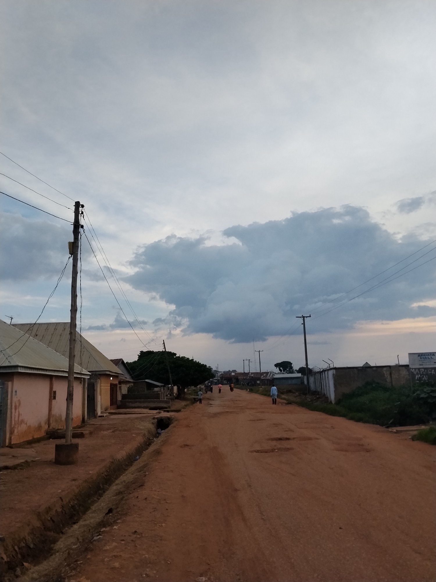 This media file captures a dirt road in Mangu Town, Nigeria, under a cloudy sky. The scene is likely set in the late afternoon or early evening, given the diffused light and the gathering clouds which suggest an approaching storm.
The road itself is unpaved, with visible potholes and a drainage ditch running alongside it. On the left, a row of single-story buildings with corrugated metal roofs lines the street. One building, painted a pale pink, has the number "11" visible on its exterior. A wooden utility pole with wires stretches across the scene, supporting electrical infrastructure. Further down the road, more buildings and trees can be seen.
Several individuals are present on the road, appearing to be walking. One person in a light blue garment is walking away from the camera. Two other figures are visible further in the distance. The presence of people and the everyday buildings suggest this is a residential or commercial area within the town.
On the right side of the road, a concrete wall with a wire fence on top marks a boundary. Behind the wall, overgrown vegetation is visible. A sign is affixed to the wall, partially obscured by the foliage and shadows. Visible text on the sign includes "AGUATA HALL" and "MARIFA MSU". The overall atmosphere is one of a quiet, possibly humid, day in a rural or semi-urban Nigerian setting. The cloudy sky and the unpaved road create a somewhat muted and earthy color palette.