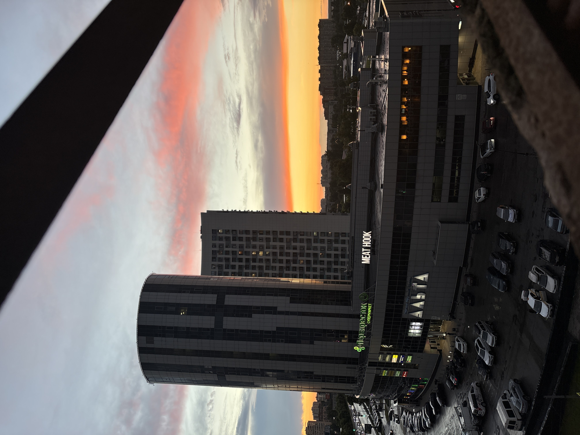 The image captures a twilight scene in Mytishchi, Russia, featuring modern architecture against a colorful sky. In the foreground, a dark, textured surface, possibly a railing or balcony edge, obscures the lower right corner. Dominating the midground is a complex of buildings, including a distinctive cylindrical skyscraper with a facade of alternating dark and light vertical panels, punctuated by numerous windows. To its right stands a taller, rectangular building, also clad in panels and filled with illuminated windows.
The lower section of the complex is a commercial building with a glass facade displaying multiple signs, including "ПЯТЕРОЧКА СУПЕРМАРКЕТ" (Pyaterochka Supermarket) and "MEAT HOOK". Another large illuminated sign reads "АДЭНТА". A wet parking lot is visible in front of the commercial building, filled with a variety of parked cars, including sedans, SUVs, and vans. The ground appears reflective, suggesting recent rain.
Beyond the immediate buildings, the urban landscape of Mytishchi stretches out, with residential buildings and trees visible under a sky painted with hues of orange, yellow, and pink from the setting sun, interspersed with gray clouds. Traffic lights and vehicles on a street are discernible on the left side of the image. The overall impression is a view of a contemporary urban environment at dusk.