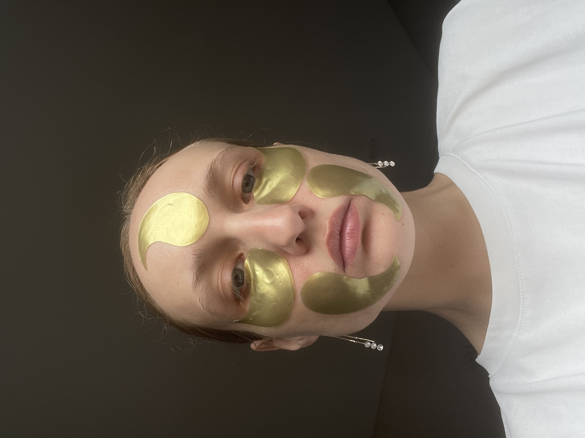 A fair-skinned person is seen from a low angle, facing the camera. They have gold-colored patches applied to their face: one on the forehead, one under each eye, and one on the upper lip. They are wearing a white T-shirt. There is a black background behind them. A decorative hair clip with small jewels is visible near the side of their face. There is no information that suggests this image was taken in the city of Подшивалово, Russia.