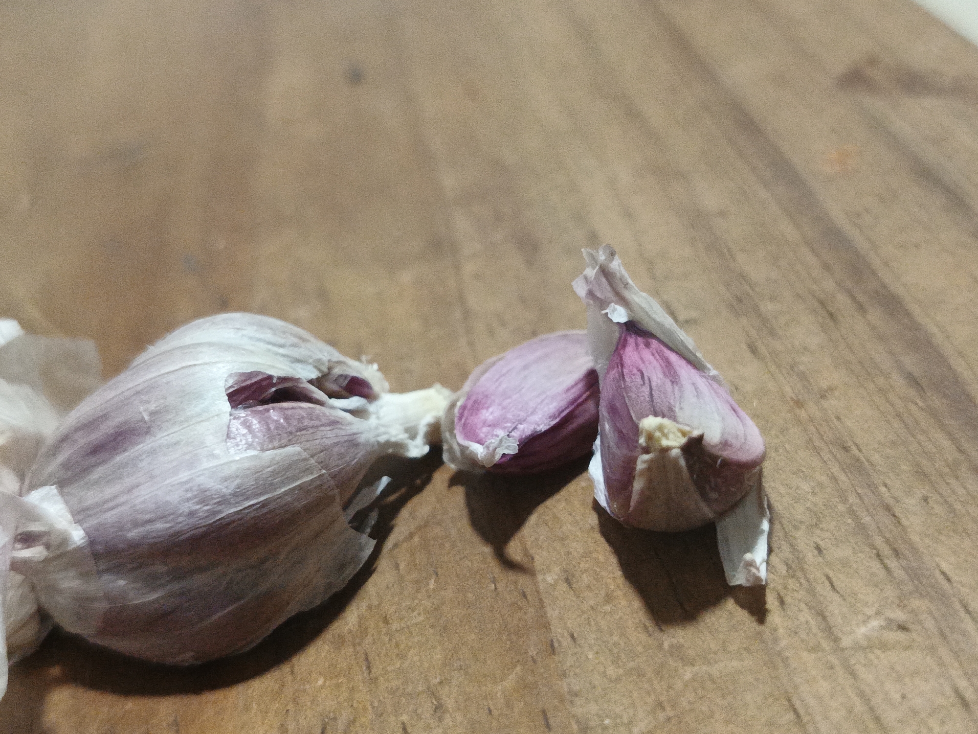 The image shows a head of garlic and two loose cloves on a wooden surface. The head of garlic is partially opened, revealing some of the purple cloves inside. The two loose cloves are also purple. The wooden surface appears to be a cutting board or tabletop. The lighting is somewhat dim, and there are no other objects or people visible in the frame. The location of Los Robles, Venezuela is not discernible from the image.