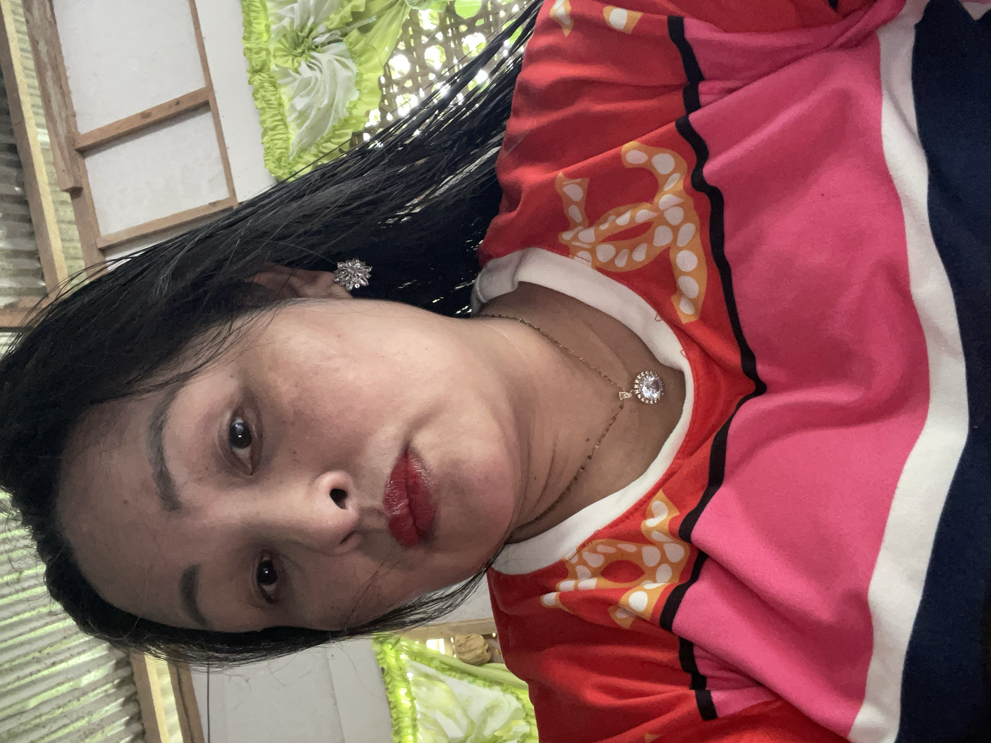 A woman with dark hair, red lipstick, and jewelry is in Hijo, Philippines. She wears a top with red, pink, black, white, and yellow patterns. The background includes what appears to be a window with a green and white curtain or covering.