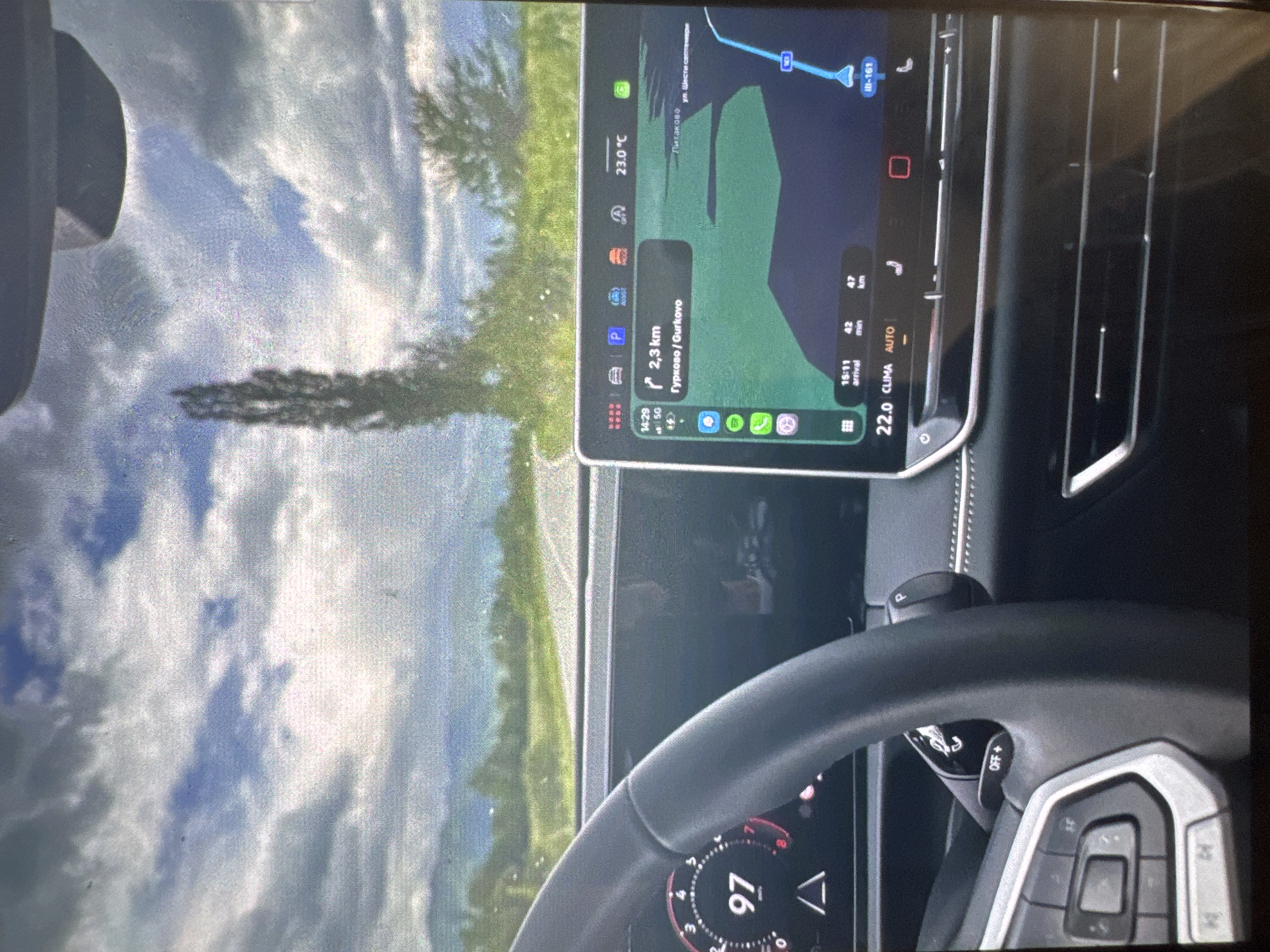 The image displays a first-person perspective from within a modern vehicle, looking forward through the windshield while simultaneously showing interior dashboard elements. On the left, a portion of a black steering wheel with silver controls is visible, partially obscuring the driver's digital display, which prominently shows a speed of "97 km/h" and a hazard warning symbol.
To the right, a large central infotainment touchscreen displays detailed navigation information. The screen indicates the current time as 14:29 and a next turn in 2.3 km towards "Гурково / Gurkovo". The map shows a route over land and potentially water, with smaller text labels including "Литаково" and "ул. Шести септември", and indicates the route is along road "III-161". Estimated arrival time is 15:11, with 42 minutes and 47 km remaining. The top bar of the screen shows various vehicle status icons, including an ambient temperature of 23.0 °C. The bottom section of the screen shows climate control settings at "22.0 CLIMA AUTO", along with seat heater icons. Below the screen, horizontal air vents are visible across the dashboard.
The exterior view through the windshield reveals a two-lane asphalt road extending into the distance through a rural landscape. Green foliage lines both sides of the road, with a tall, slender tree prominently positioned on the right side. The sky above is partly cloudy, with visible patches of blue interspersed with grey and white clouds.
This scene is presented as occurring in the city of Vratsa, Bulgaria, although the navigation system on the screen indicates destinations and road numbers (such as Gurkovo and Litakovo, and road III-161) located in other regions of Bulgaria.
