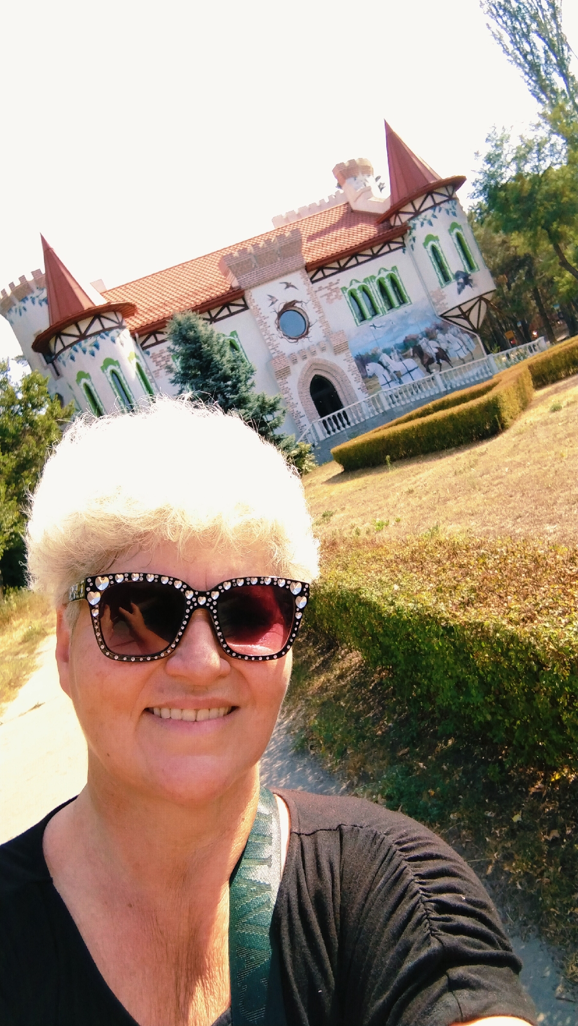 A medium shot captures a woman with short, white, curly hair and large, heart-shaped sunglasses adorned with rhinestones. She is smiling broadly and wearing a black t-shirt, with a green, patterned strap visible over her shoulder. Behind her, a whimsical, colorful building designed to resemble a castle is prominent. The castle features red conical roofs, arched windows with green trim, and a mural depicting horses and riders. The building is situated in an outdoor, park-like setting with dry grass, manicured hedges, and trees. The scene is bathed in bright sunlight. This is likely the "Fairy Tale Castle" located in the city of Horishni Plavni, Ukraine.