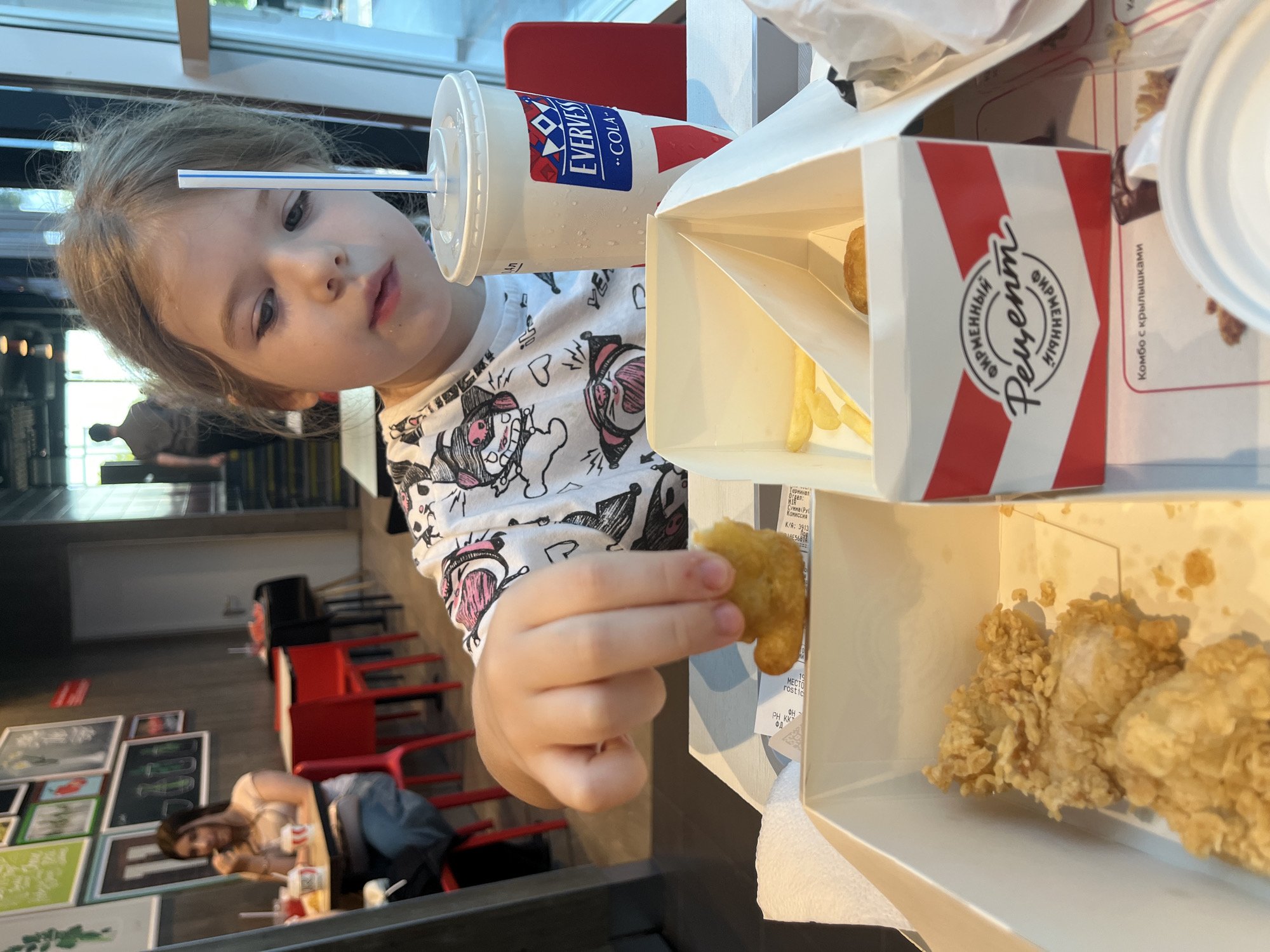 A young Caucasian girl with blonde hair is holding a chicken nugget over an open white cardboard box containing french fries and another chicken nugget, in what appears to be a fast-food restaurant. The girl is wearing a white t-shirt with a cartoon print. To her right is a large white disposable cup with a blue and red logo that reads "EVERVESS COLA." The cup has a white lid and a blue and white striped straw. In the background, another person is seated at a table with red chairs. The restaurant has dark gray walls with framed pictures and a large window. The overall lighting suggests an indoor, daytime setting. The restaurant is likely located in Sochi, Russia, as indicated by the text on the box, which appears to be in Russian and reads "Фирменный Рецепт" (Firm Favorite Recipe).
