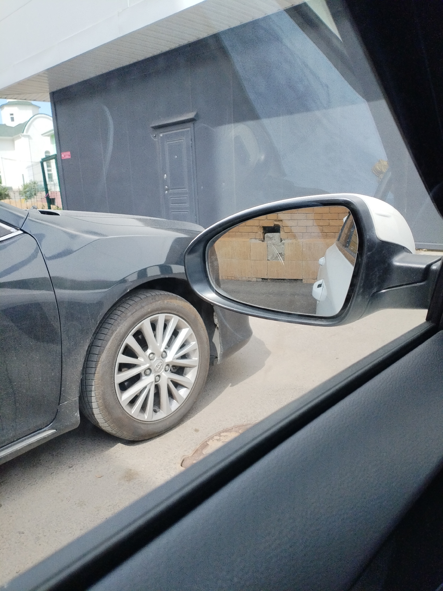 An exterior, eye-level, medium shot captures the side of a dark gray sedan parked on an asphalt surface next to a building. The car's front fender and wheel are visible on the left. A side-view mirror on the car's passenger side reflects another white vehicle and a brick wall. The building behind the sedan is dark gray with a white, horizontally paneled overhang. A white building with a green roof is partially visible in the background. The scene appears to be a parking area in a city.