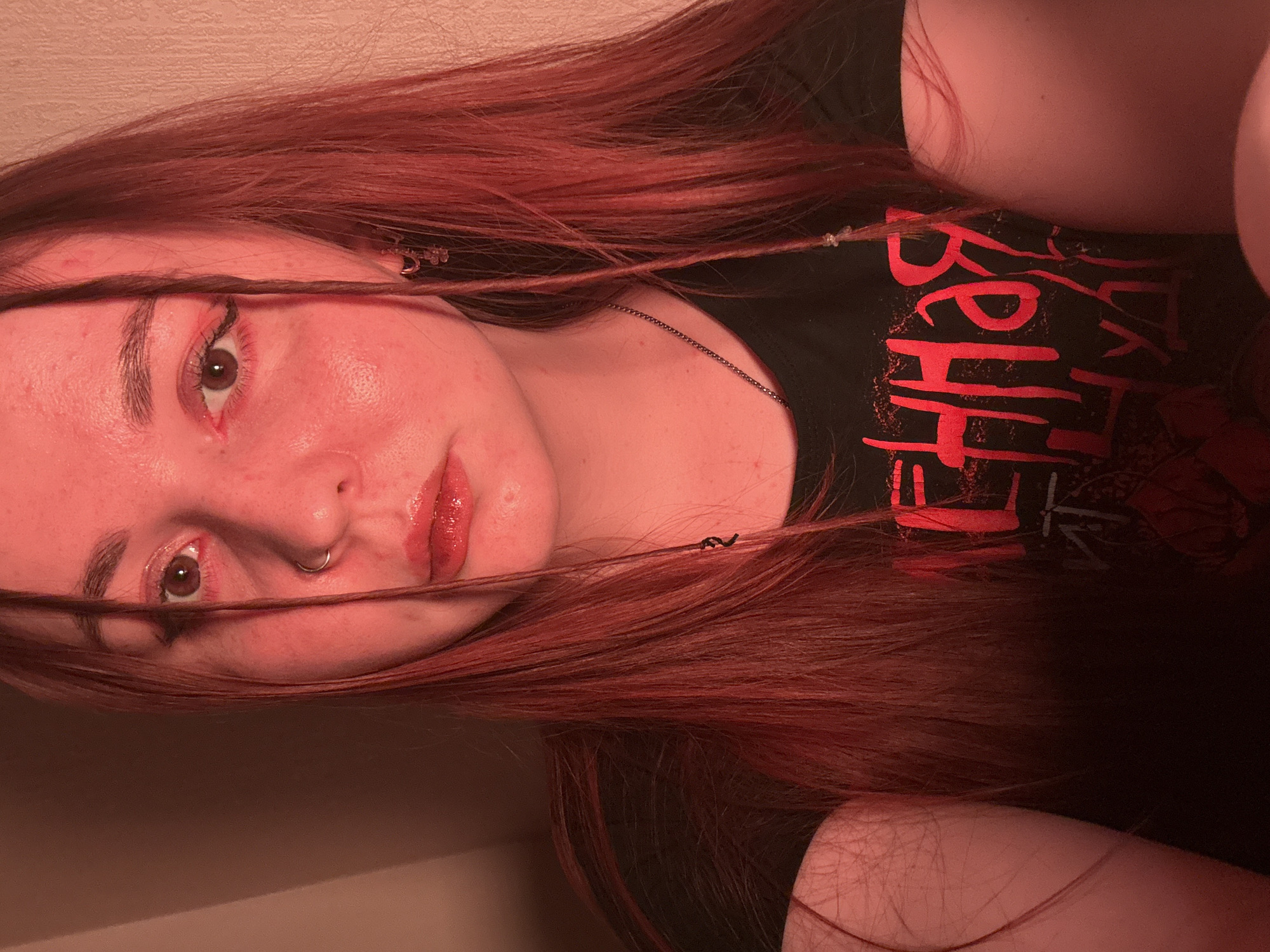A close-up selfie of a person with long, red hair. The person has fair skin and brown eyes. They are wearing a black t-shirt with red lettering. A thin chain is visible around their neck. The person has a nose ring and is wearing red lipstick and what appears to be red eye makeup. Two strands of hair are styled with small clear decorative beads. The background is a light beige or off-white color. The scene does not appear to be in Izhevsk, Russia.