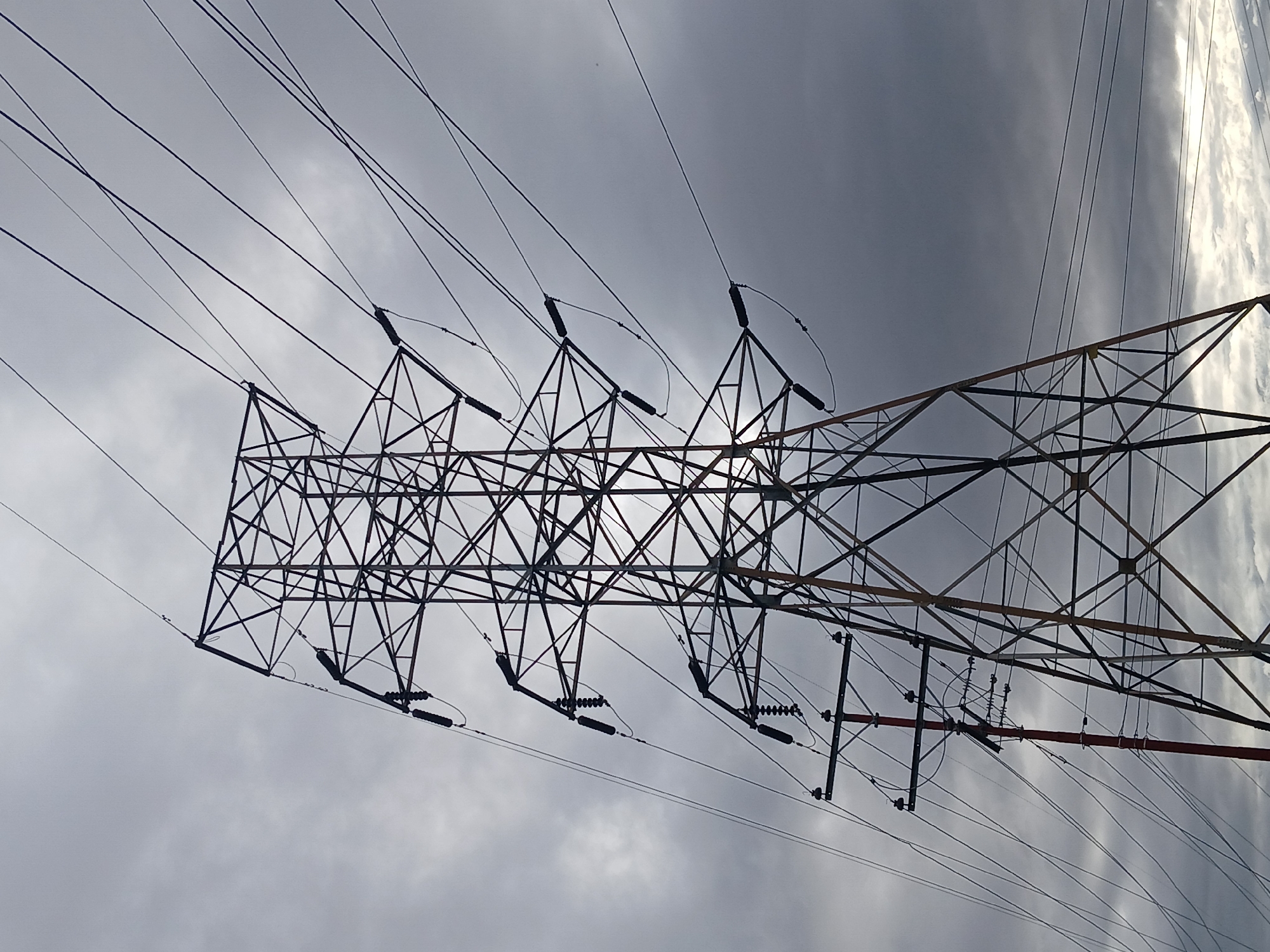 A large, dark-colored lattice-steel electrical transmission tower stands prominently, angled from the lower left to the upper right of the frame, silhouetted against an overcast sky. Numerous high-voltage power lines extend diagonally and horizontally from the tower, supported by multiple strings of dark, cylindrical insulators. To the lower-left of the main tower, a narrower, reddish-brown utility pole with crossarms and smaller insulators is visible, also supporting multiple wires. The sky is predominantly grey with varied cloud formations, including brighter, less dense clouds towards the lower right. No individuals or vehicles are visible. The scene depicts an electricity infrastructure landscape in Ciudad Guayana, Venezuela.