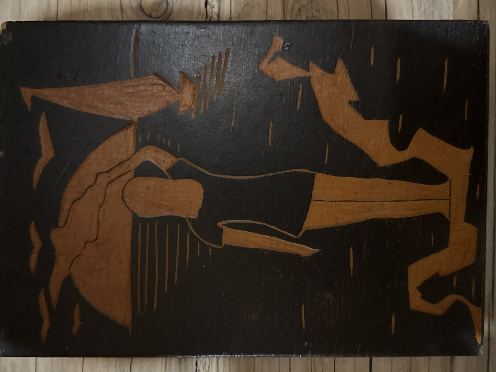 A rectangular wooden plaque with a dark background and lighter-colored carvings depicts a stylized scene. A reclining figure, possibly female, is positioned horizontally across the center of the plaque. To the left of the figure, a container or object resembling a vessel is shown. To the right of the figure, there is an abstract or stylized shape that may represent an action or another object. Vertical lines are scattered throughout the background, possibly representing rain or some other environmental effect. The style of the art is simplistic and potentially folk art. Маза, Russia is not discernable from the image.
