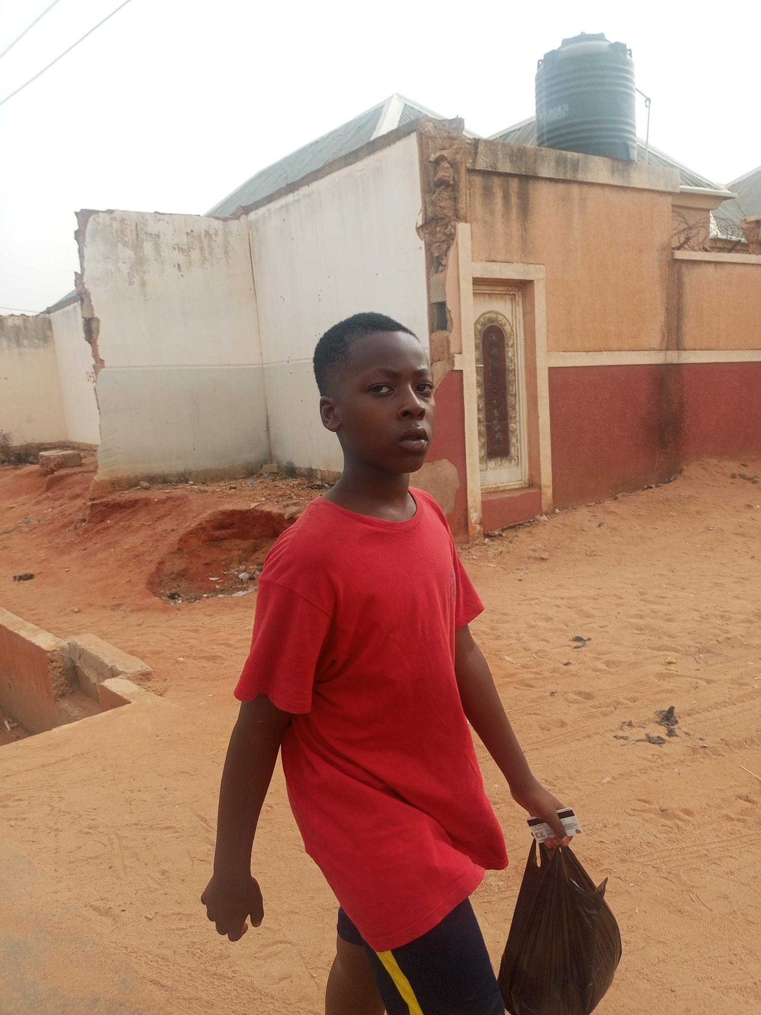 This outdoor photograph, set in Tempure, Nigeria, features a young, dark-skinned person, likely a boy, standing slightly off-center to the left. He looks directly at the camera with a neutral and calm expression. He is wearing a plain, loose-fitting red t-shirt and dark shorts with a yellow stripe running down the side. In his right hand, he holds a black plastic bag, with a white rectangular card, displaying the "Mastercard" logo, partially visible clutched between his fingers and the bag handle.
The ground is predominantly reddish-brown sand or dirt, suggesting an unpaved area. In the background, there is a building structure with walls painted in shades of white, beige, and reddish-brown. The building shows signs of wear or incomplete construction, with some sections appearing damaged or exposed, particularly on the left side. A decorative door or window frame with an arched top is visible on the building's right side. On the roof of the structure, a large, black cylindrical water tank is clearly visible against a bright, overcast sky, with the word "RAGEN" faintly legible on its side. The diffused lighting suggests it is daytime, possibly late morning or early afternoon, under a cloudy or hazy sky, as there are no distinct shadows. The scene portrays a mundane moment, with the individual pausing for the photograph in an everyday setting.
