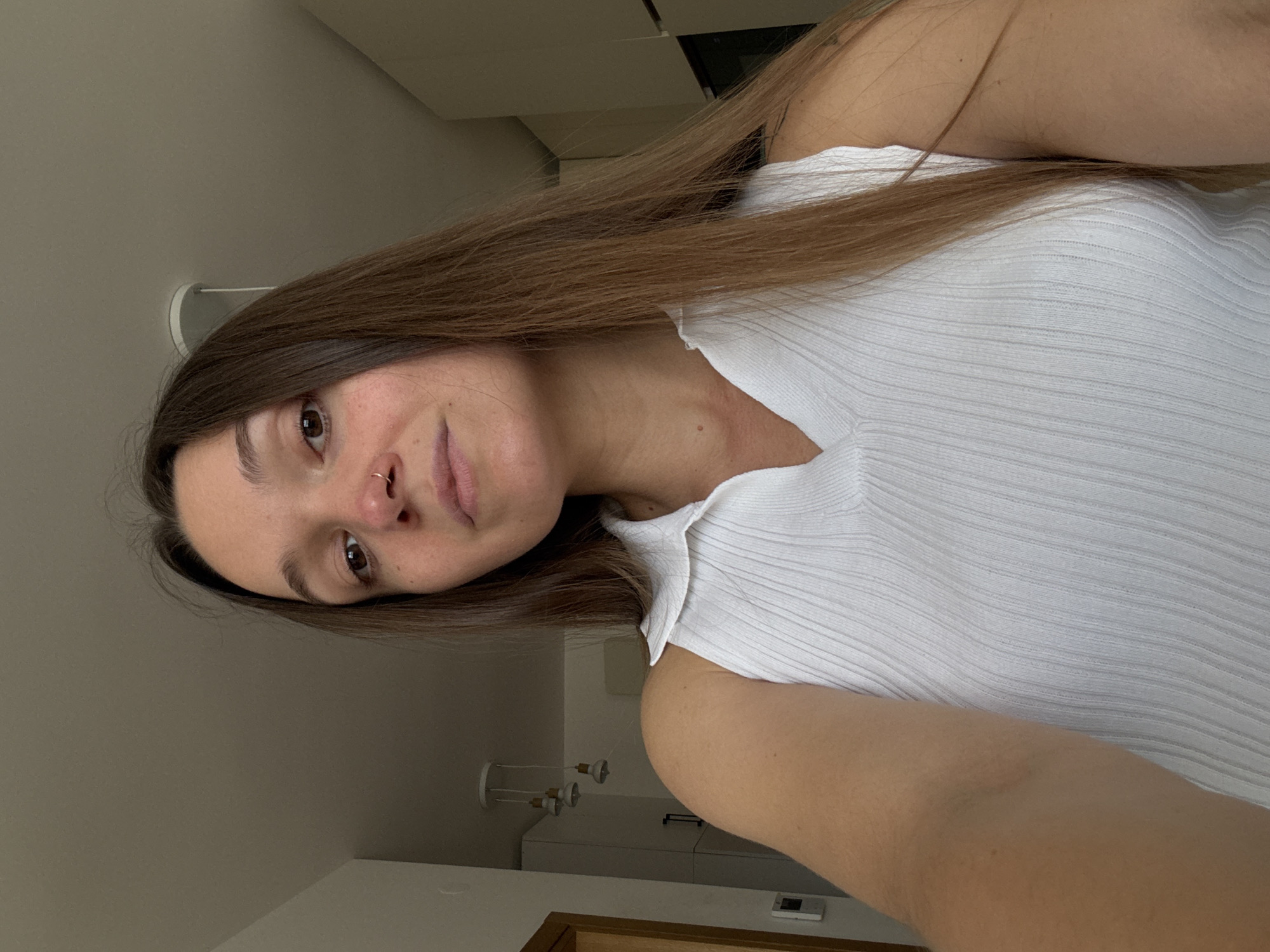 The image is a close-up, selfie-style shot of a young woman with long, brown hair. She is wearing a white, ribbed, sleeveless top with a collar and a V-neck. Her head is slightly tilted, and she is looking directly at the camera. She has a nose ring. The background appears to be an indoor domestic setting, possibly an apartment, with a ceiling and a light fixture visible. There are no discernible elements in the image that indicate the location is the city of Boreti, Montenegro.