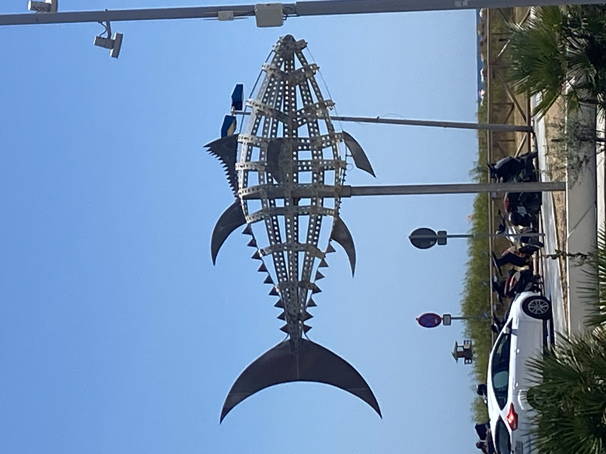 The image depicts a large, metallic fish sculpture mounted on poles, dominating the foreground under a clear blue sky. The sculpture, constructed from a skeletal framework of metal bars and circular perforated panels, resembles a tuna with prominent dorsal and tail fins. Behind the sculpture, a white car is parked on a street, with several motorcycles clustered nearby. In the background, a lifeguard tower stands amidst greenery, with a wooden fence bordering the area. Beyond the fence, a beach can be seen with colorful umbrellas and people scattered along the shoreline. The scene appears to be a sunny day in Conil de la Frontera, Spain, likely near the coast, given the presence of the beach and lifeguard tower.