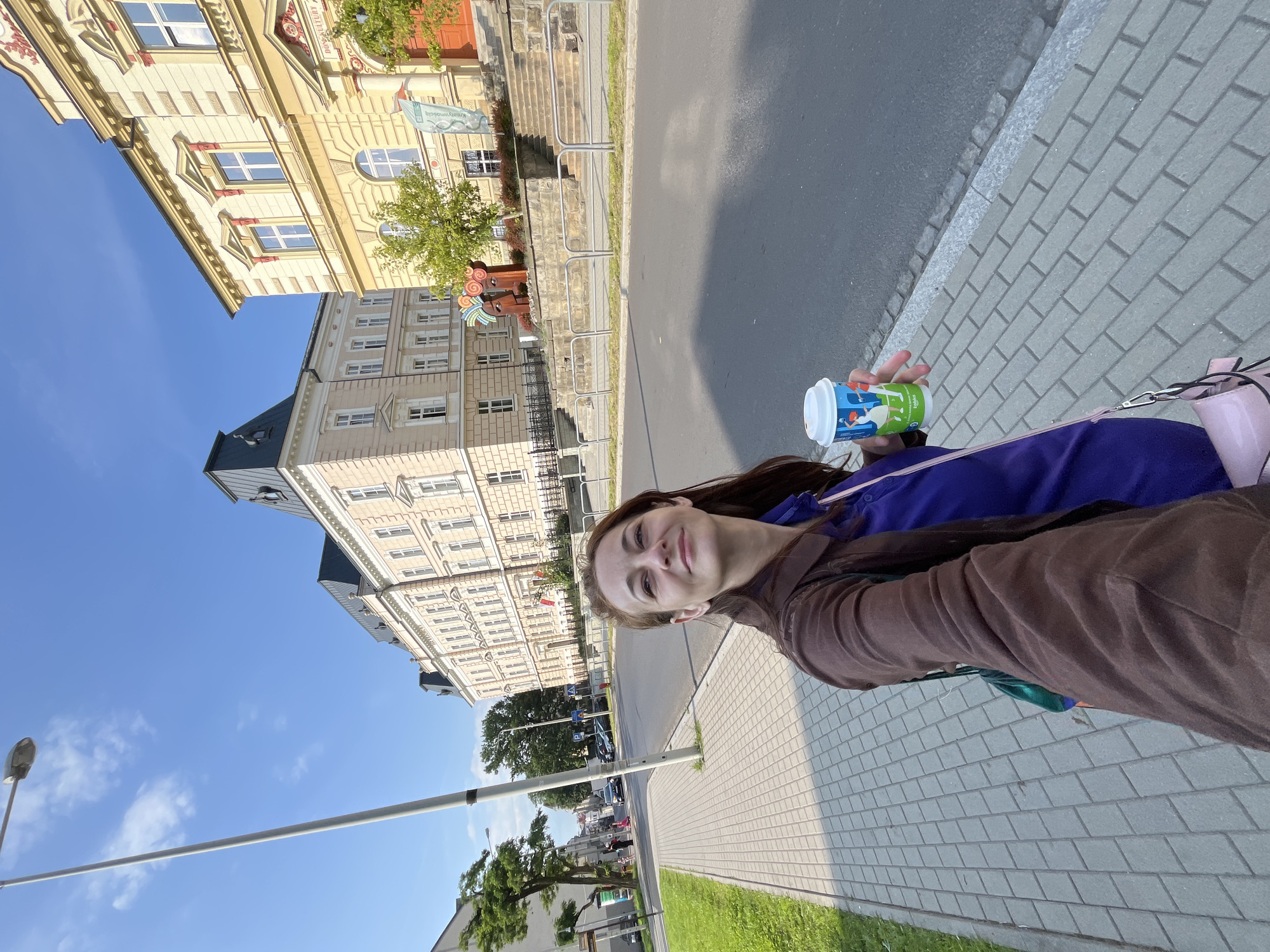In this outdoor, medium shot selfie, a woman with long brown hair, wearing a brown jacket over a purple shirt and a pink purse, walks on a sidewalk next to a street. She holds a blue and white disposable coffee cup with cartoon drawings of swans. The woman is looking up and to her right, smiling. The background features a street with parked cars, buildings with light-colored facades and dark roofs, and a bright blue sky with scattered clouds. A tall, grey street lamp stands on the left side of the image. The sidewalk is paved with grey rectangular tiles, and a strip of green grass is visible between the sidewalk and the street. In the middle ground, a sculpture of two stylized heads with colorful swirls adorns the area in front of a building.
