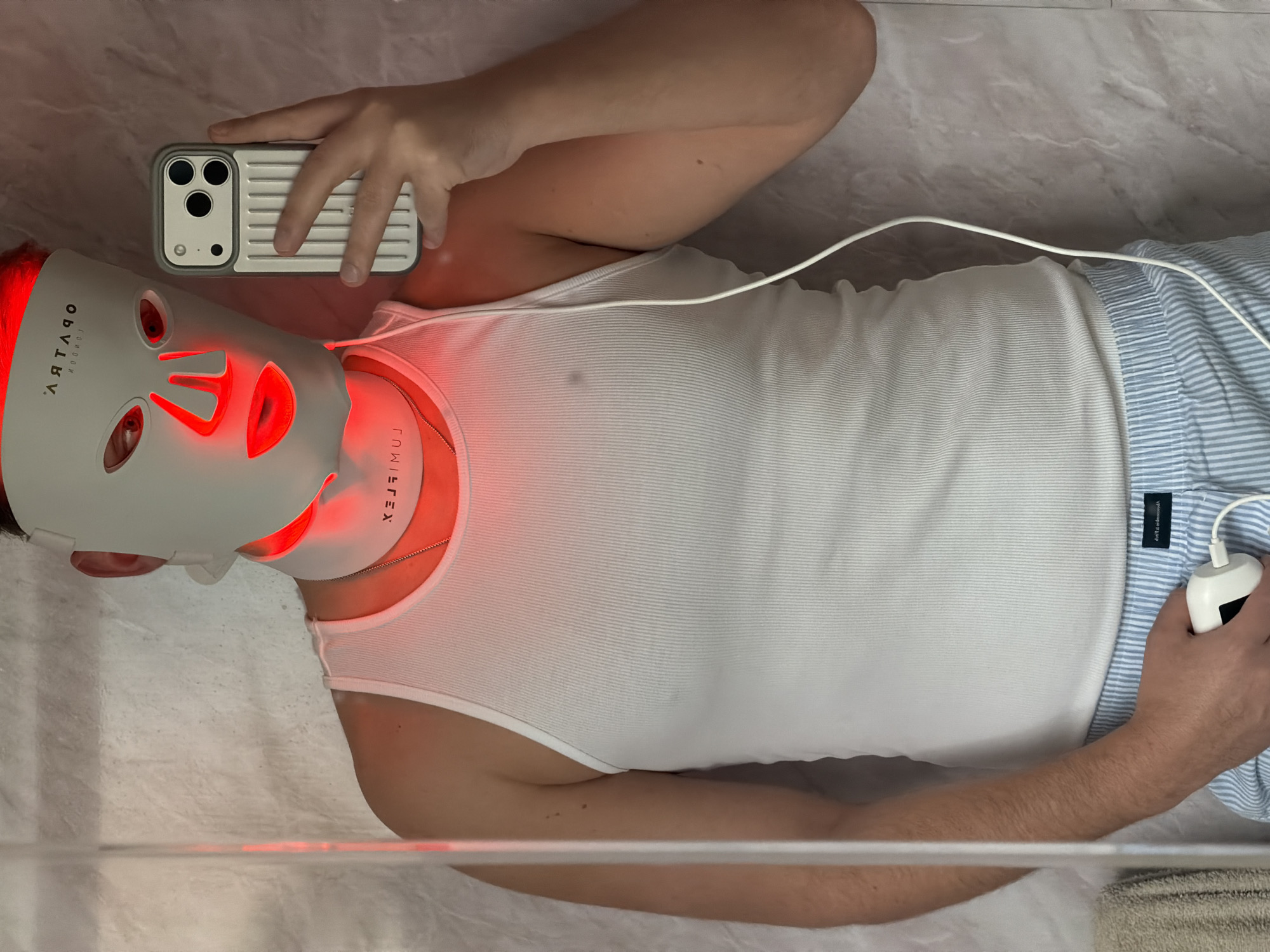 A person lies on their back, wearing a white tank top and blue striped pajama bottoms, with a white LED face mask emitting red light. They are holding a white smartphone in one hand and a white control device for the mask in the other. The person appears to be indoors, lying on a white surface with a marbled pattern. The mask is labeled "OPTIMV". The city and exact location are not discernible from the image.
