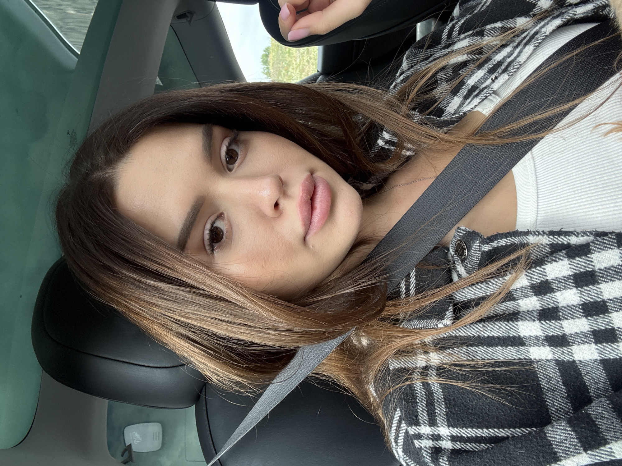 A young woman with long, dark brown hair and light makeup is sitting in the driver's seat of a car. She is wearing a white ribbed tank top and a black and white plaid shirt. A gray seatbelt is diagonally across her chest. Her right hand is resting on her lap, with her fingers slightly curled. Her left hand is partially visible, extended towards the right side of the frame. The interior of the car appears to be black leather. Through the windshield, a blurry outdoor scene of green foliage and a cloudy sky is visible. The image is a close-up, focusing on the woman's face and upper body. The location is identified as Prague, Czechia.