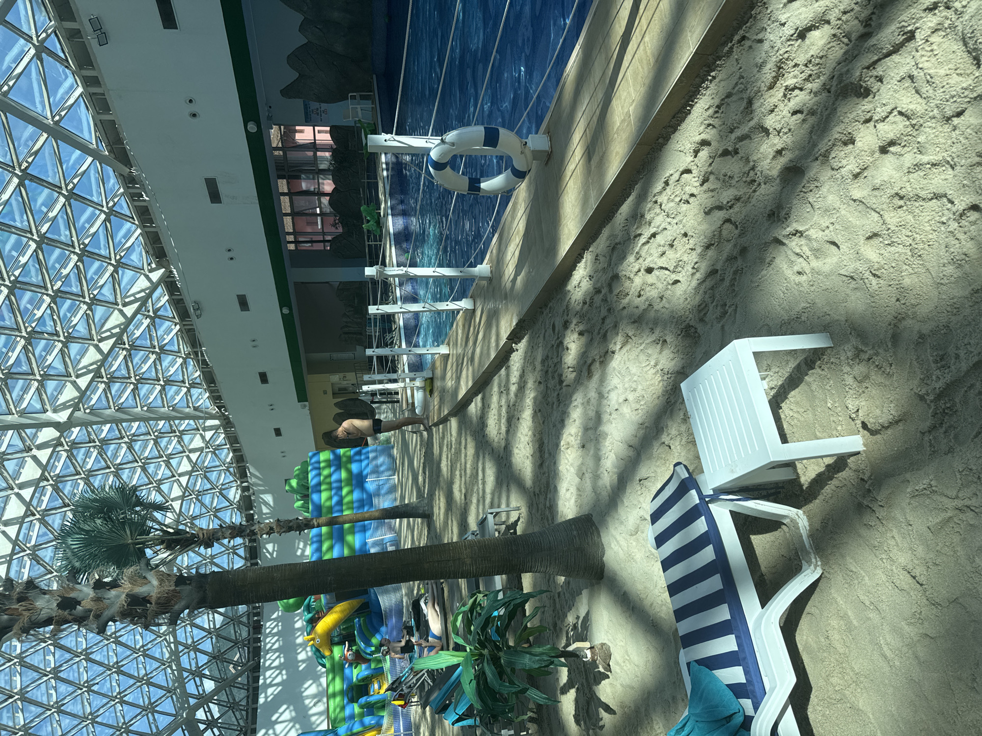 This image depicts an indoor water park in Estosadok, Russia, under a large geodesic dome with a triangular glass roof. The scene features a sand-covered area with several lounge chairs, one of which has a blue and white striped cushion and a turquoise towel draped over it. A small white plastic table sits next to the lounge chair. In the background, a large swimming pool with clear blue water is visible, bordered by a wooden walkway and white rope fencing. A life preserver, white with blue stripes, is mounted on a post near the pool. Palm trees, both real and artificial, are scattered throughout the sandy area, adding to a tropical theme. A large inflatable water play structure with bright green, blue, and yellow elements is also present. Several individuals are visible in the scene: one person stands near the pool, looking down at their phone; another person is seated on a lounge chair in the sand; and two other individuals are seated further back in the sandy area. The overall environment suggests a leisure and recreational space designed to simulate a beach experience indoors.