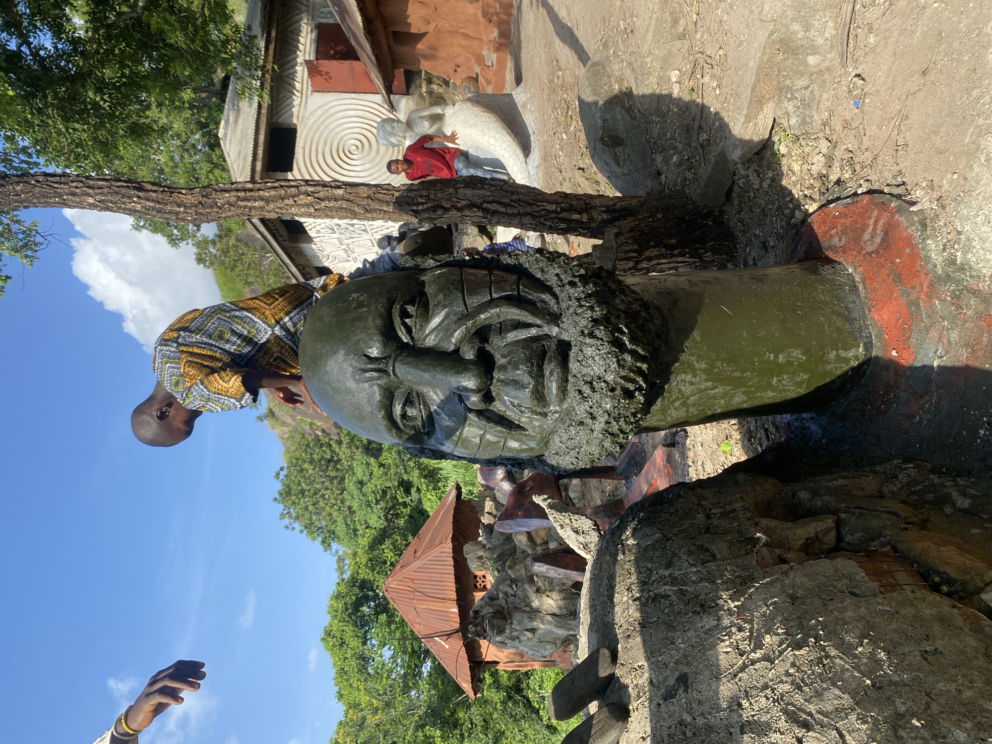 A close-up, eye-level outdoor shot captures a large, dark green, rough-textured statue of a human head with a beard in the foreground. The statue is positioned slightly off-center to the right. Behind the statue, a young boy wearing a patterned shirt is bent over, interacting with the top of the statue. To the left of the frame, an arm with a yellow bracelet reaches into the scene. In the middle ground, there are several small, rustic structures with corrugated metal roofs, along with other statues and rock formations. A boy in a red shirt is visible sitting on a mermaid statue in the background, near a tree. The environment is lush and green with trees and foliage, under a clear blue sky with some scattered clouds. The ground is dirt and gravel, with large rocks scattered around. The overall scene appears to be an outdoor art installation or cultural site.