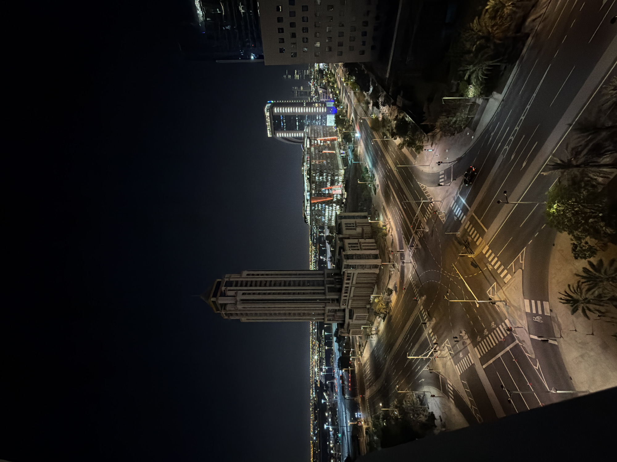 A night aerial view captures a city intersection with a tall, rectangular skyscraper prominently featured on the left. The skyscraper has a stepped pyramid-like crown and is illuminated by the dark sky. A complex road network with multiple lanes and pedestrian crossings dominates the foreground and midground, with traffic lights visible at the intersections. Several cars are present on the roads. In the background, other illuminated buildings of varying heights form a cityscape, with distant lights suggesting a sprawling urban environment. The scene is characterized by the contrast between the dark night sky and the artificial lights from buildings and streetlights. Palm trees are scattered throughout the landscape, adding a touch of vegetation to the urban setting.