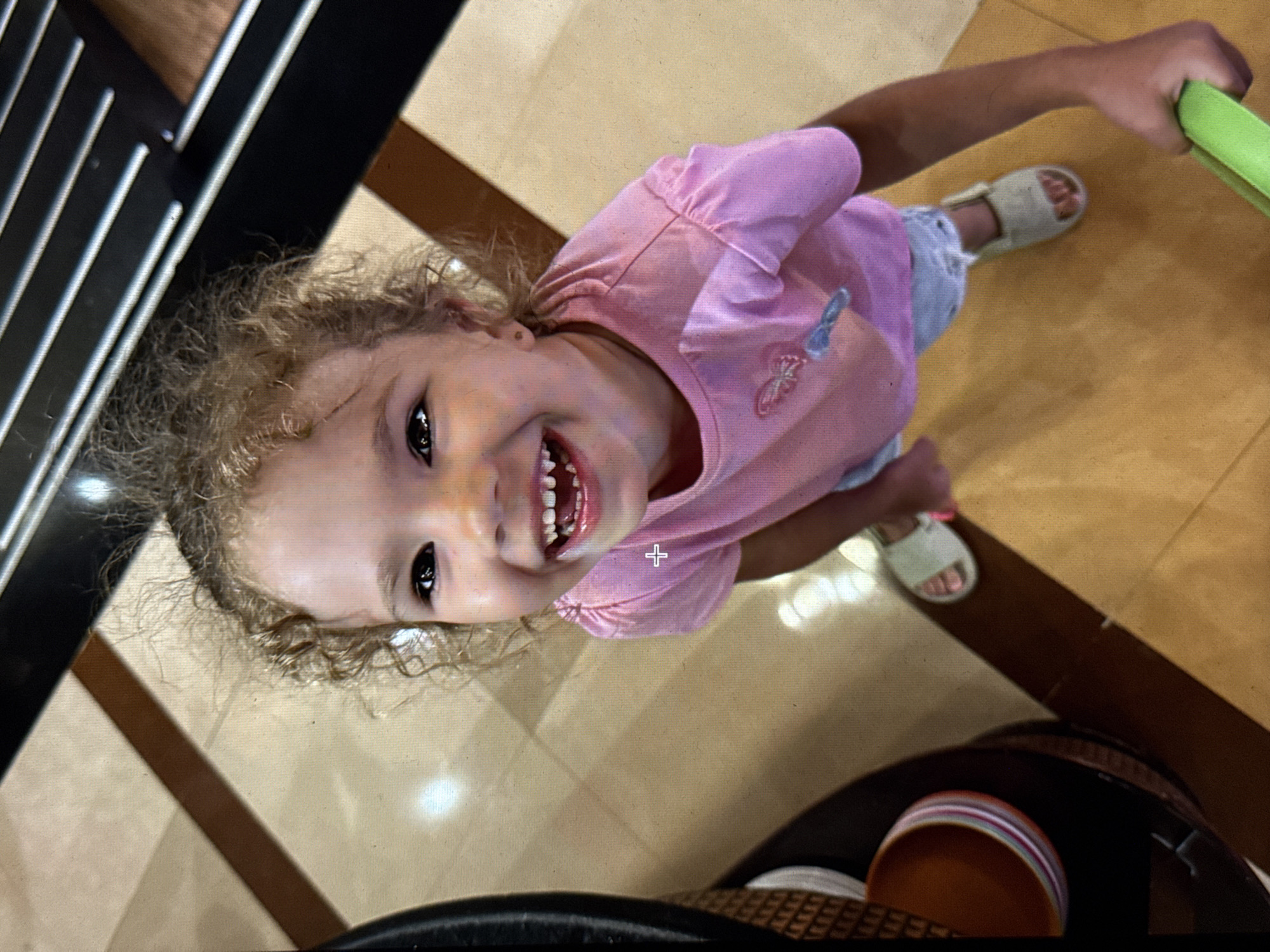 A high-angle shot captures a young child with curly blonde hair, wearing a pink shirt and light-colored shorts. She is smiling broadly, showing her teeth. The child is holding a green object in her right hand. She is wearing sandals. The background consists of a tiled floor and parts of furniture, including a dark-colored object with metal slats on the left, and a dark round object with a striped bowl on the bottom right. The location in Minsk, Belarus cannot be determined from the provided image.