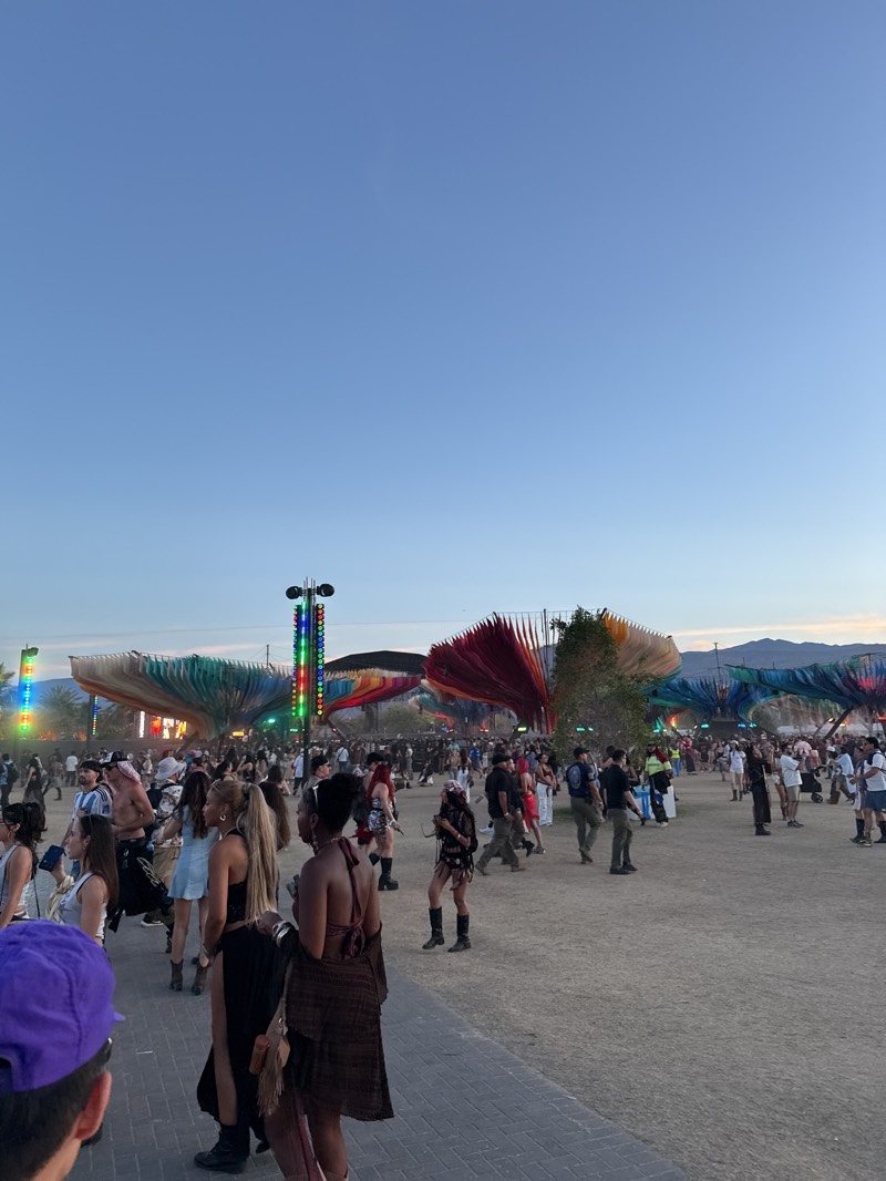 The image shows a large crowd of people at the Coachella Valley Music and Arts Festival in Indio, California, United States. The festival grounds are visible in the background, featuring colorful, large-scale art installations, trees and buildings. People are walking around, and the overall atmosphere seems lively and energetic. The sky is a clear gradient, transitioning from a bright blue. A tall light tower with multicolored lights stands close.