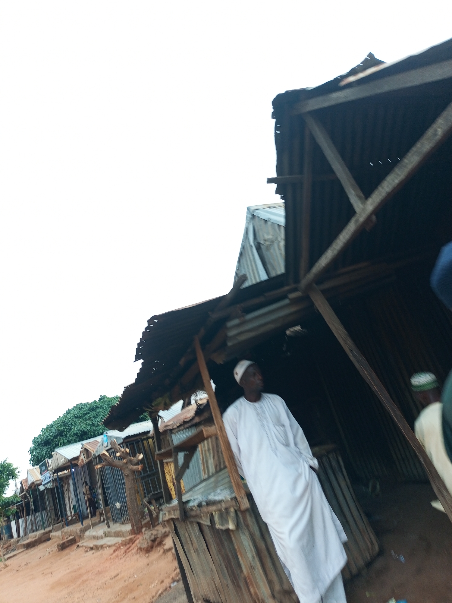 A low-angle shot captures a street scene in Tempure, Nigeria, featuring a row of small shops constructed with corrugated metal roofing and wooden structures. A man dressed in a white traditional garment and a white cap stands in the foreground, facing right. Behind him, another person in a light-colored garment and a checkered cap is visible. The ground is dusty red earth. The sky is overcast.